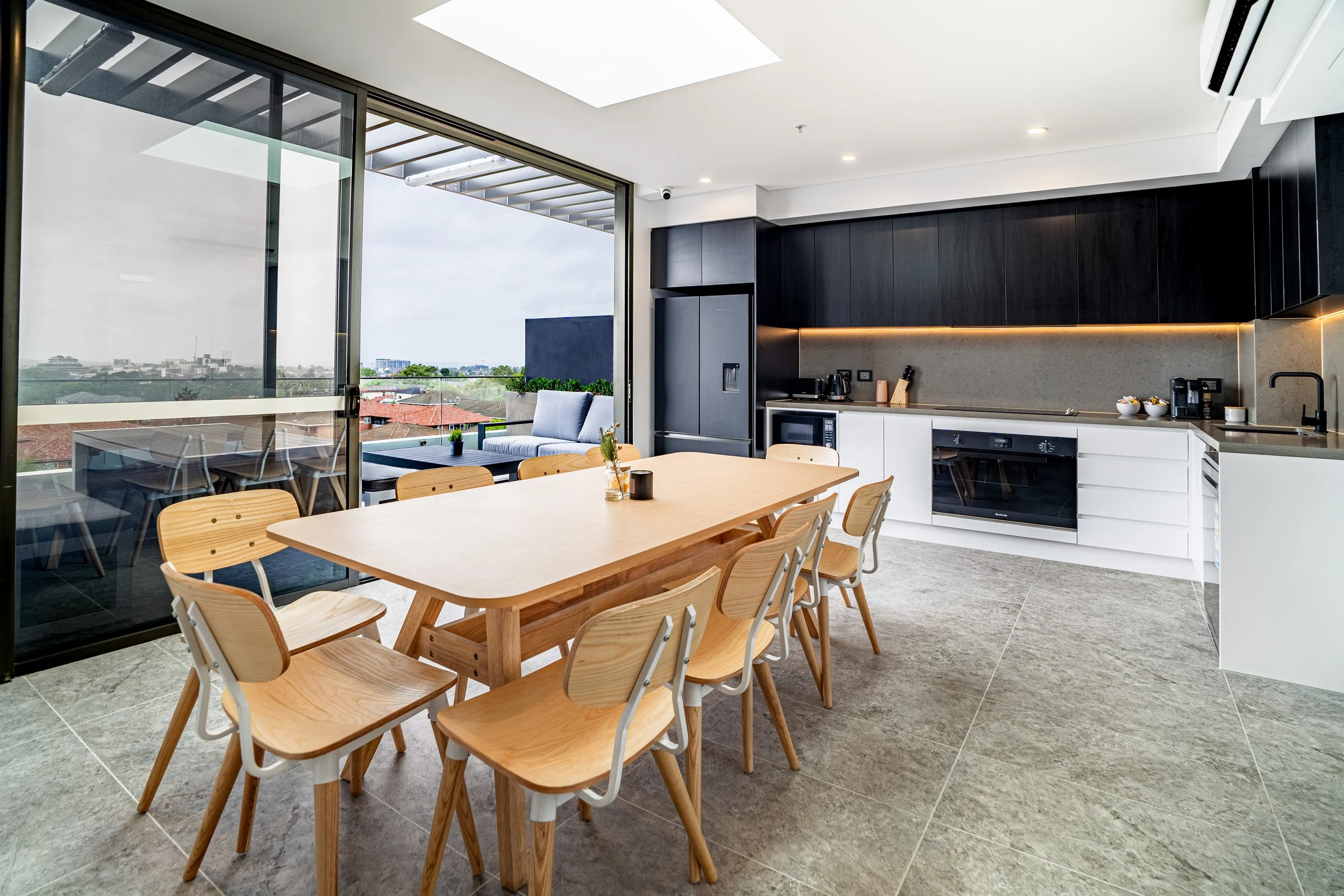 Modern kitchen with a wooden dining table and chairs, black and white cabinets, large sliding glass door leading to a balcony with outdoor seating, overlooking a cityscape with a cloudy sky.