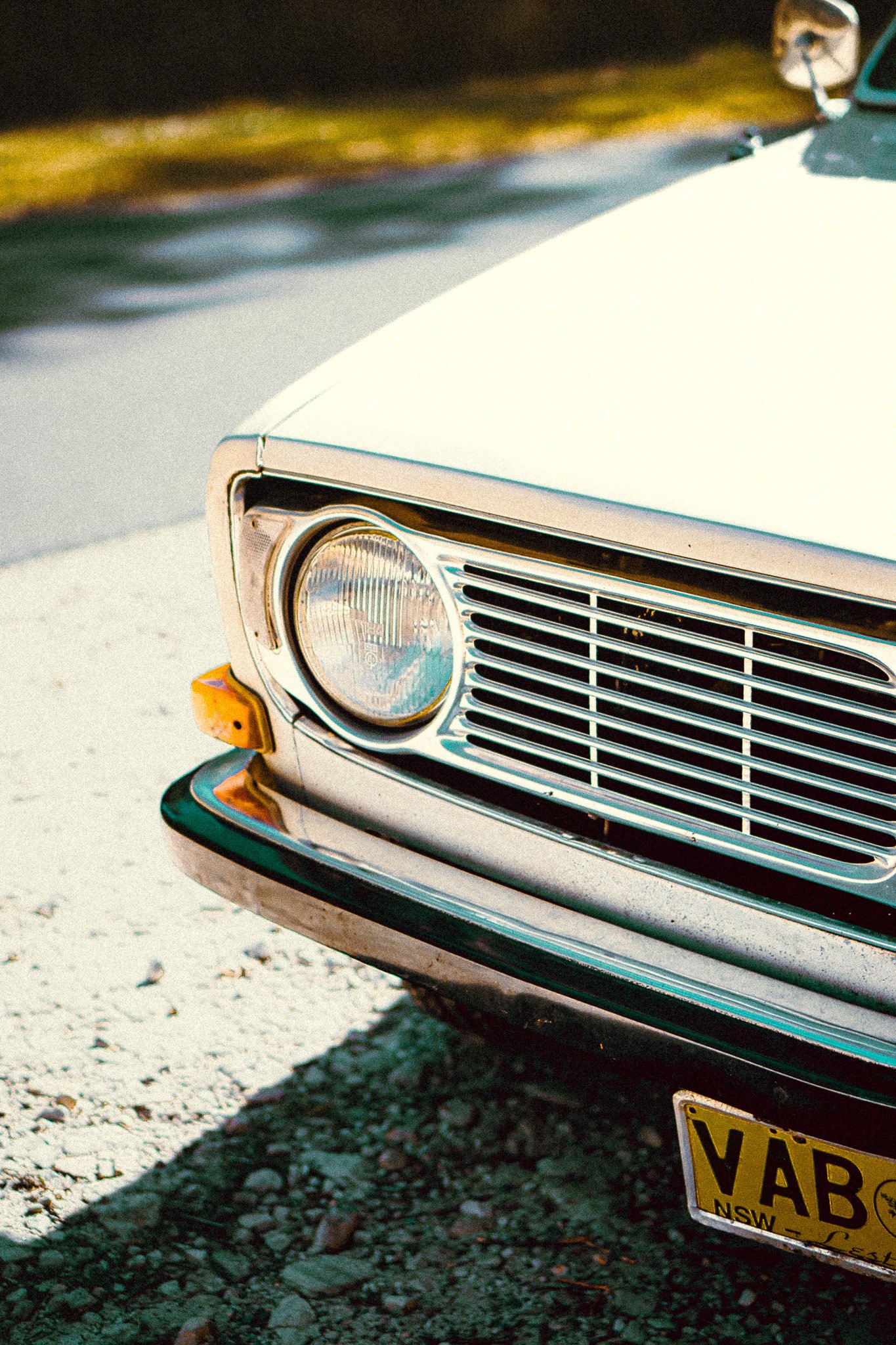 Close-up of the front of a vintage white car with a yellow license plate reading "VAB" on a gravel surface.