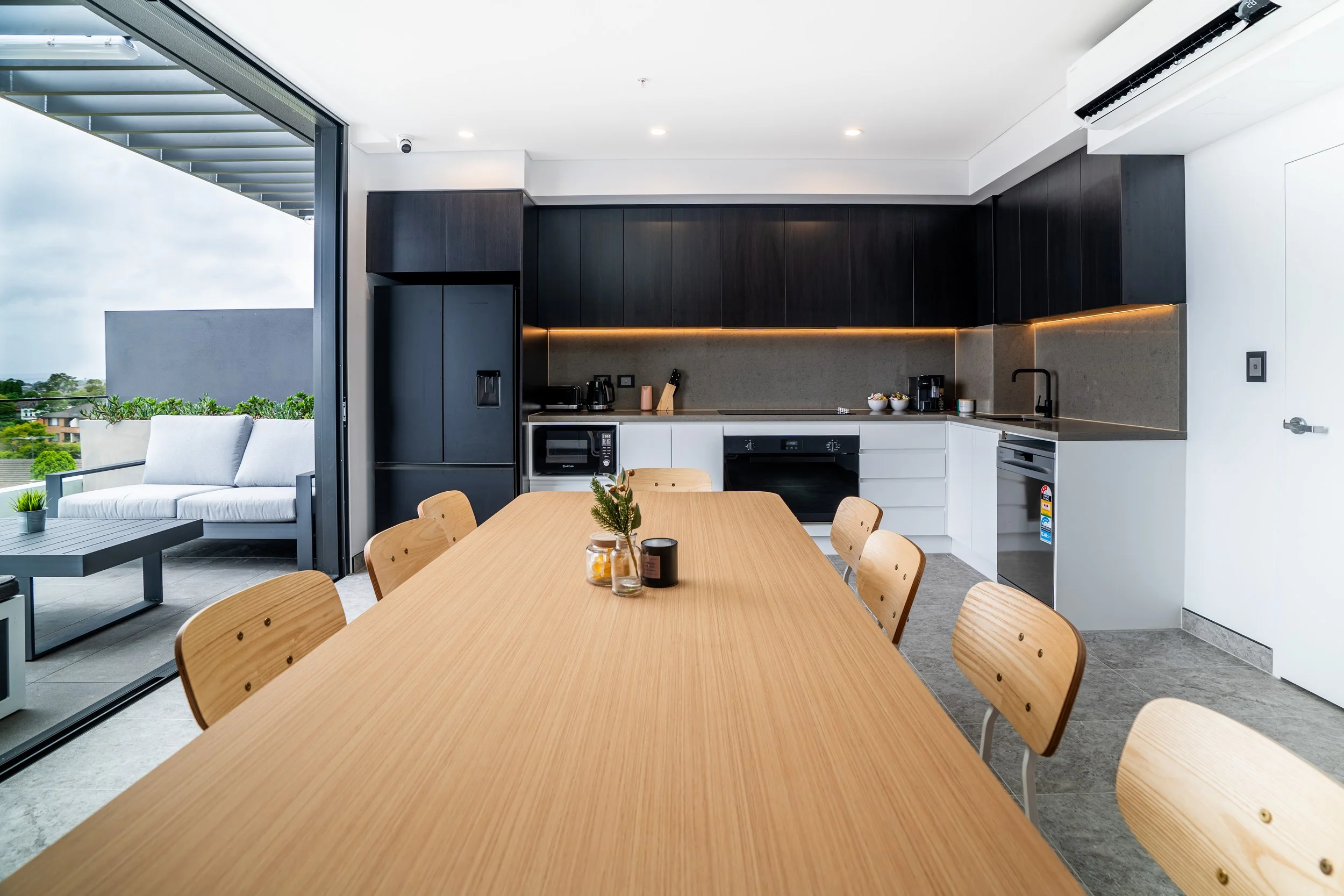 Modern kitchen and dining area with a long wooden table, black and white cabinets, and an outdoor balcony with seating.