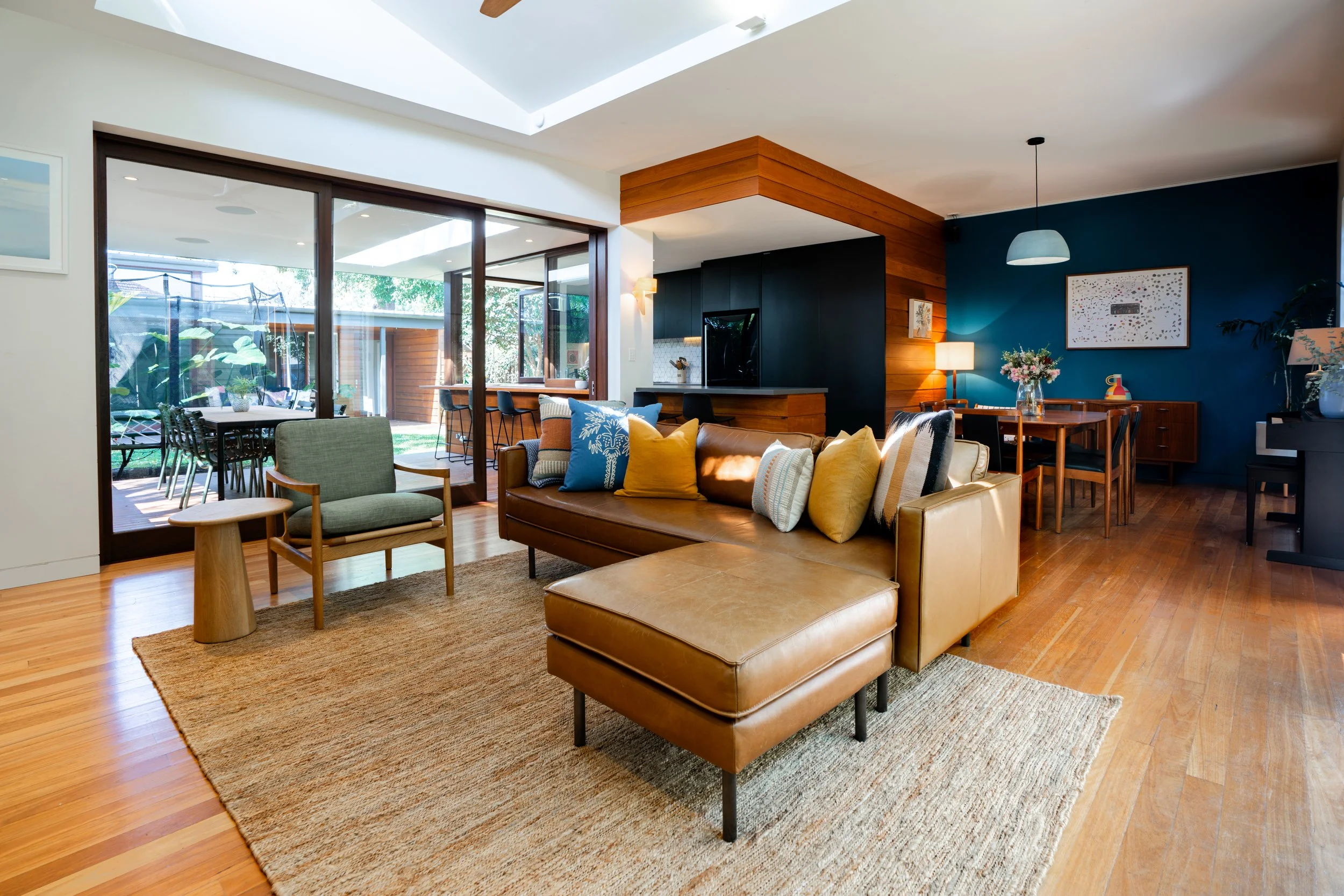 Open-concept living room and dining area with wooden floors, a leather sofa with colorful pillows, a side chair, dining table with chairs, and an outdoor patio visible through sliding glass doors.