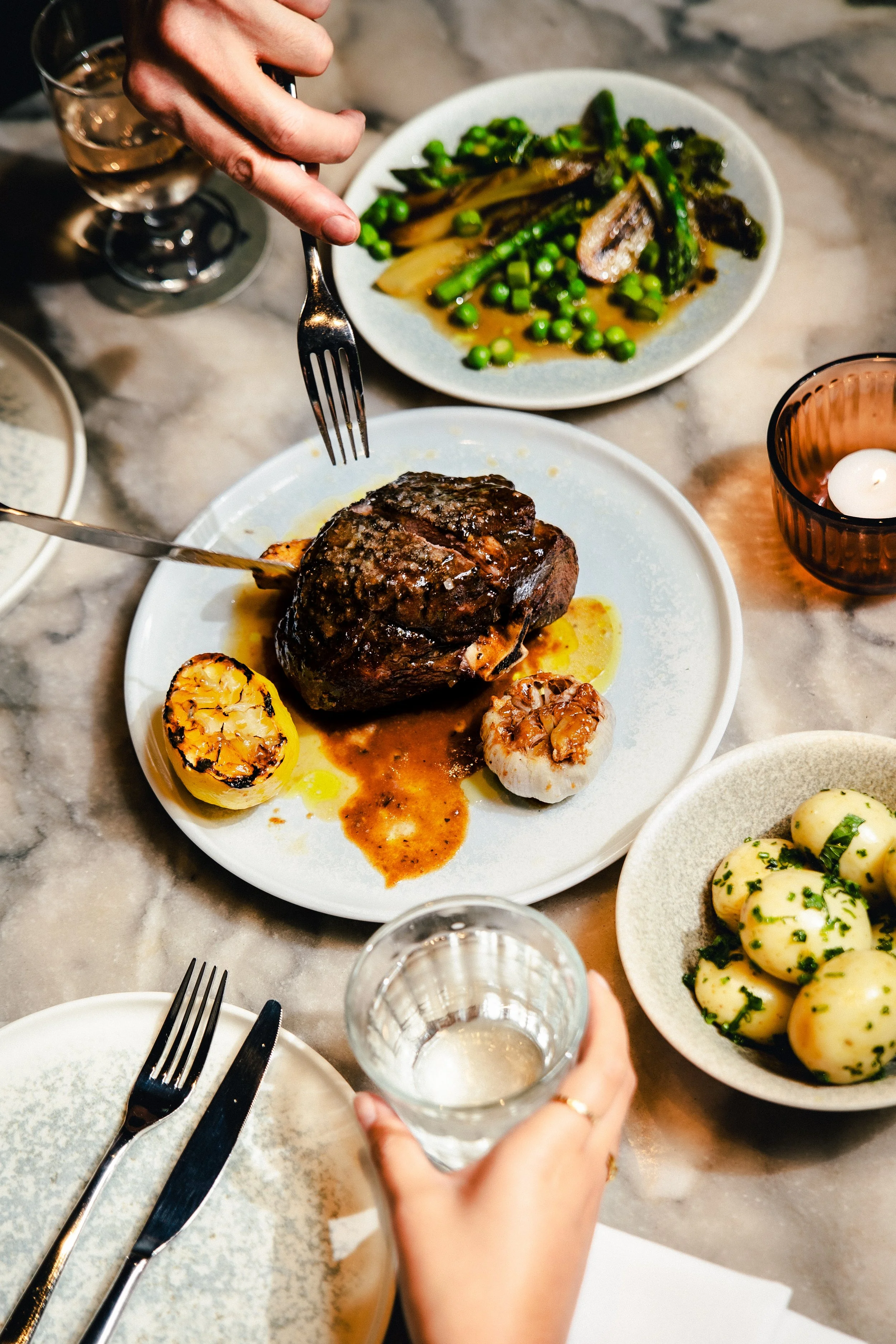 A table with a cooked steak topped with sauce, grilled garlic, and roasted garlic bulb on a white plate, accompanied by grilled vegetables, boiled potatoes with parsley, and glasses of water and white wine.