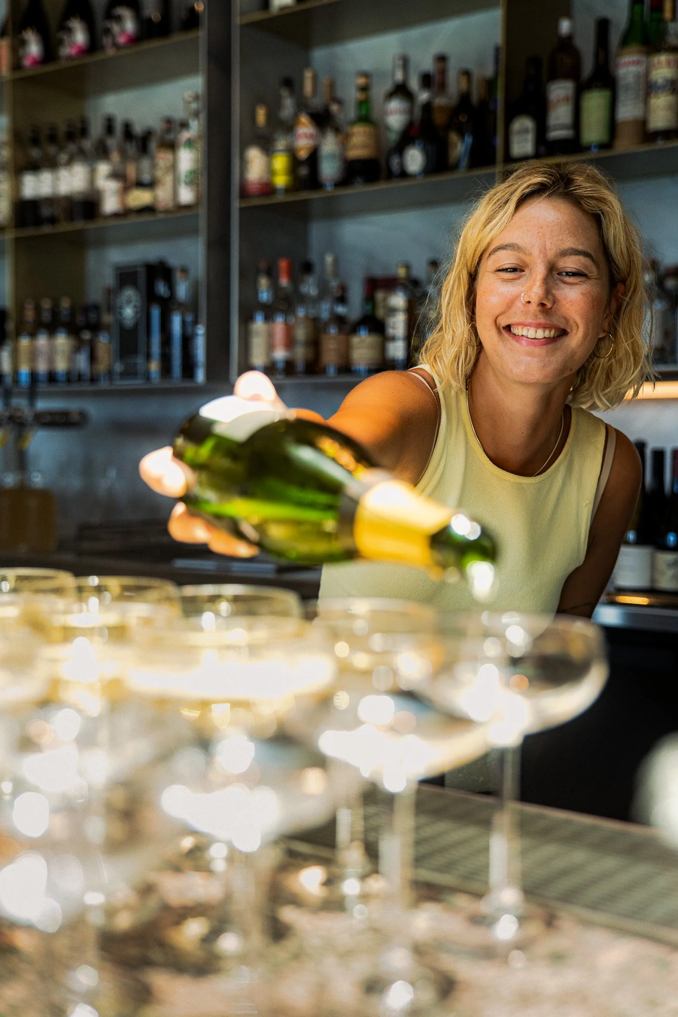 A woman with blonde hair and a yellow sleeveless top smiling while pouring drinks into glasses at a bar or restaurant. Shelves of liquor bottles are visible in the background.