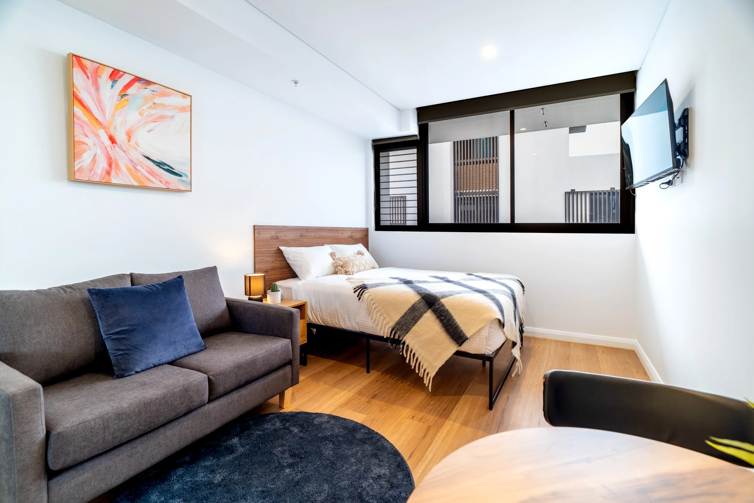 Modern studio apartment with a bed, gray sofa with a blue pillow, abstract painting on white wall, wooden floor, large window, and mounted flat-screen TV.