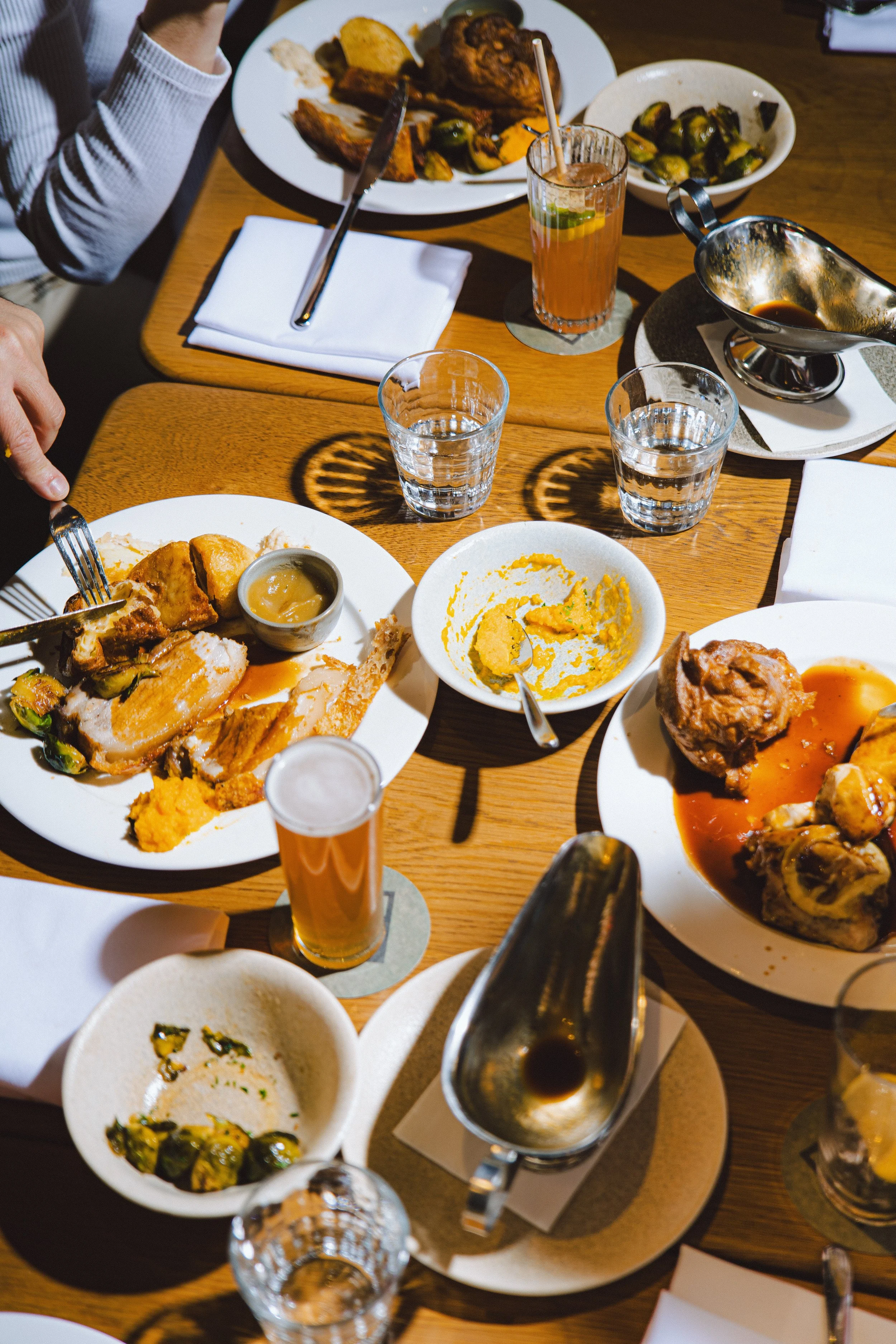 Empty plates with remnants of food, glasses of water and drinks on a wooden table, with some Brussels sprouts, mashed potatoes, roast chicken, and a gravy boat.