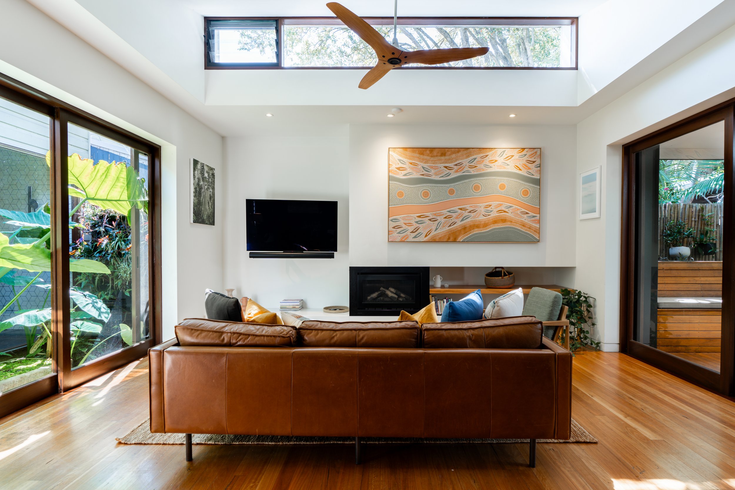 Modern living room with a brown leather sofa, a flat-screen TV on the white wall, colorful artwork above the fireplace, large windows, and sliding glass doors leading outside, with a ceiling fan and hardwood floors.