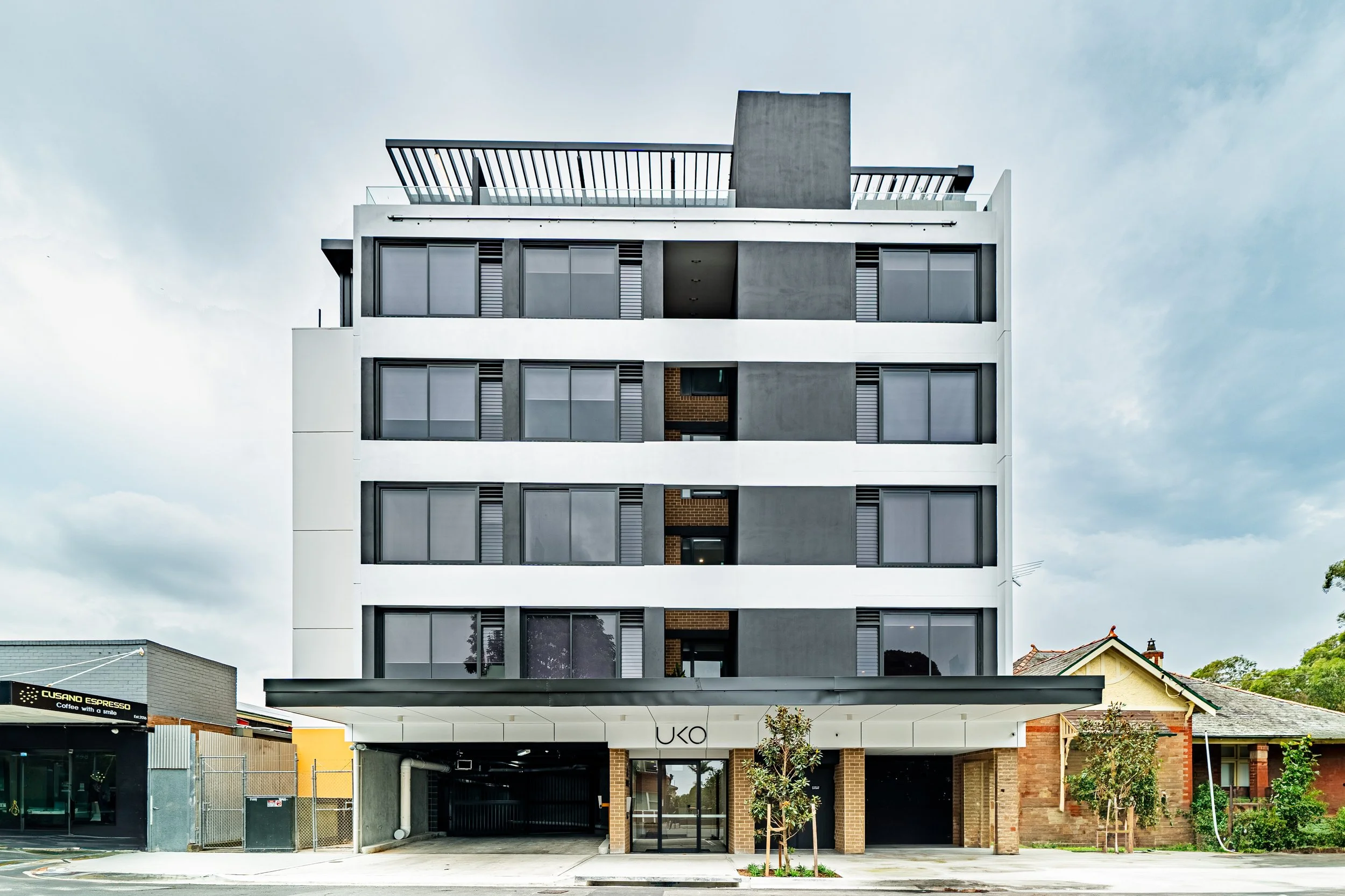 A modern multi-story apartment building with a white and gray facade, large windows, and a covered entrance labeled 'UKO.'