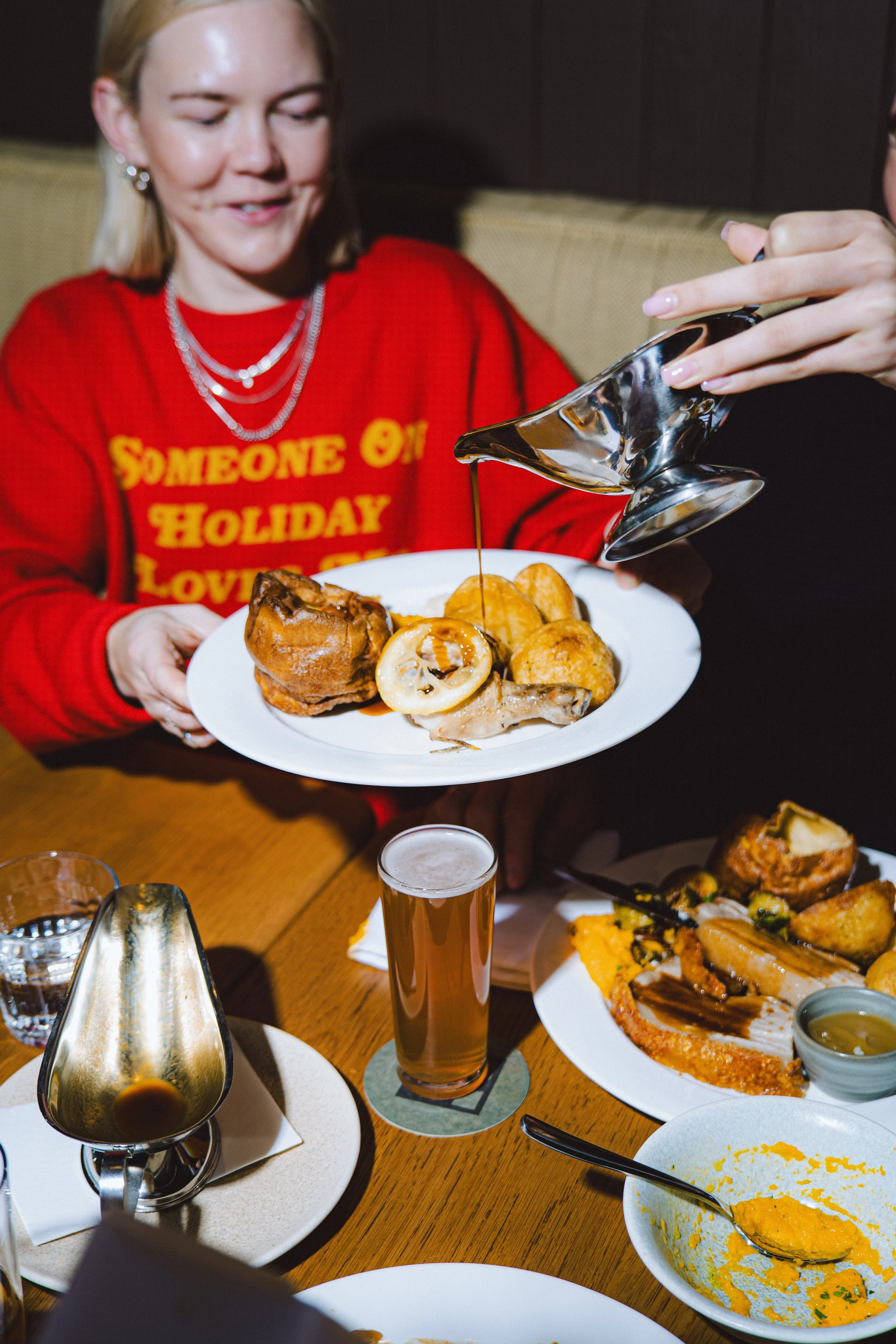 A woman in a red sweater holding a white plate with various foods, including a piece of meat, roasted potatoes, and a lemon slice. Someone is pouring gravy onto the plate. The table has drinks, including a glass of beer, and other plates of food.
