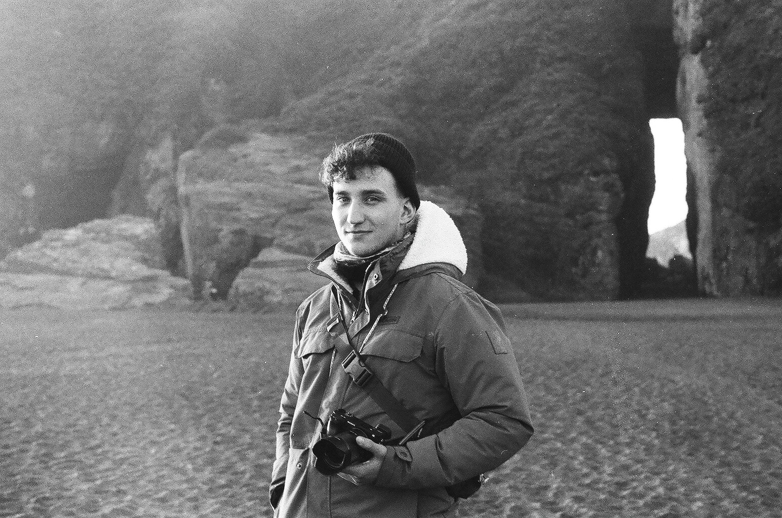 A young man holding a camera stands outdoors in front of large rock formations or cliffs, with a narrow opening or archway in the background.