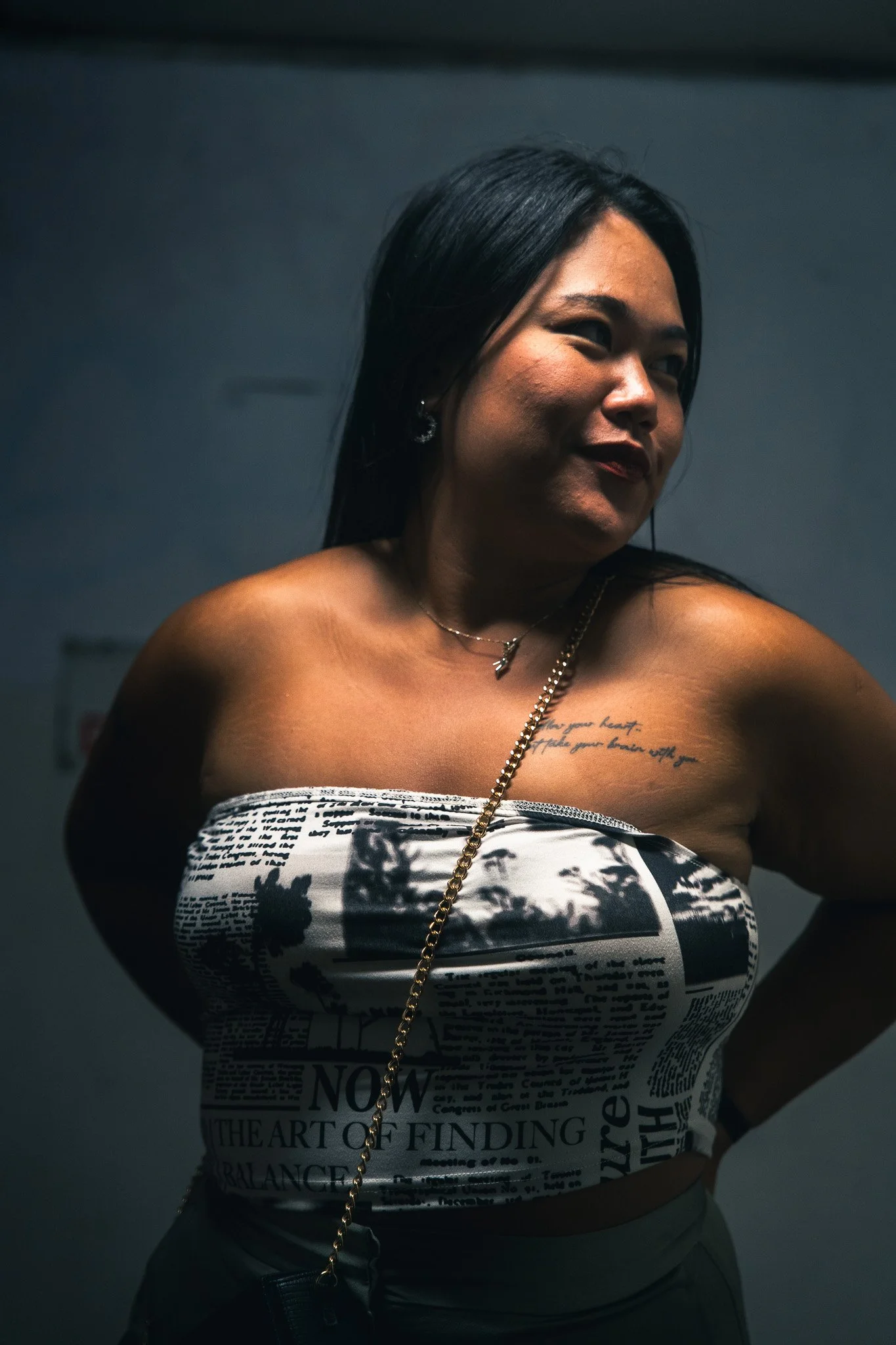 A woman with dark hair smiling and looking to her right, wearing a strapless top with newspaper print design, a necklace, and earrings, standing against a dark background.