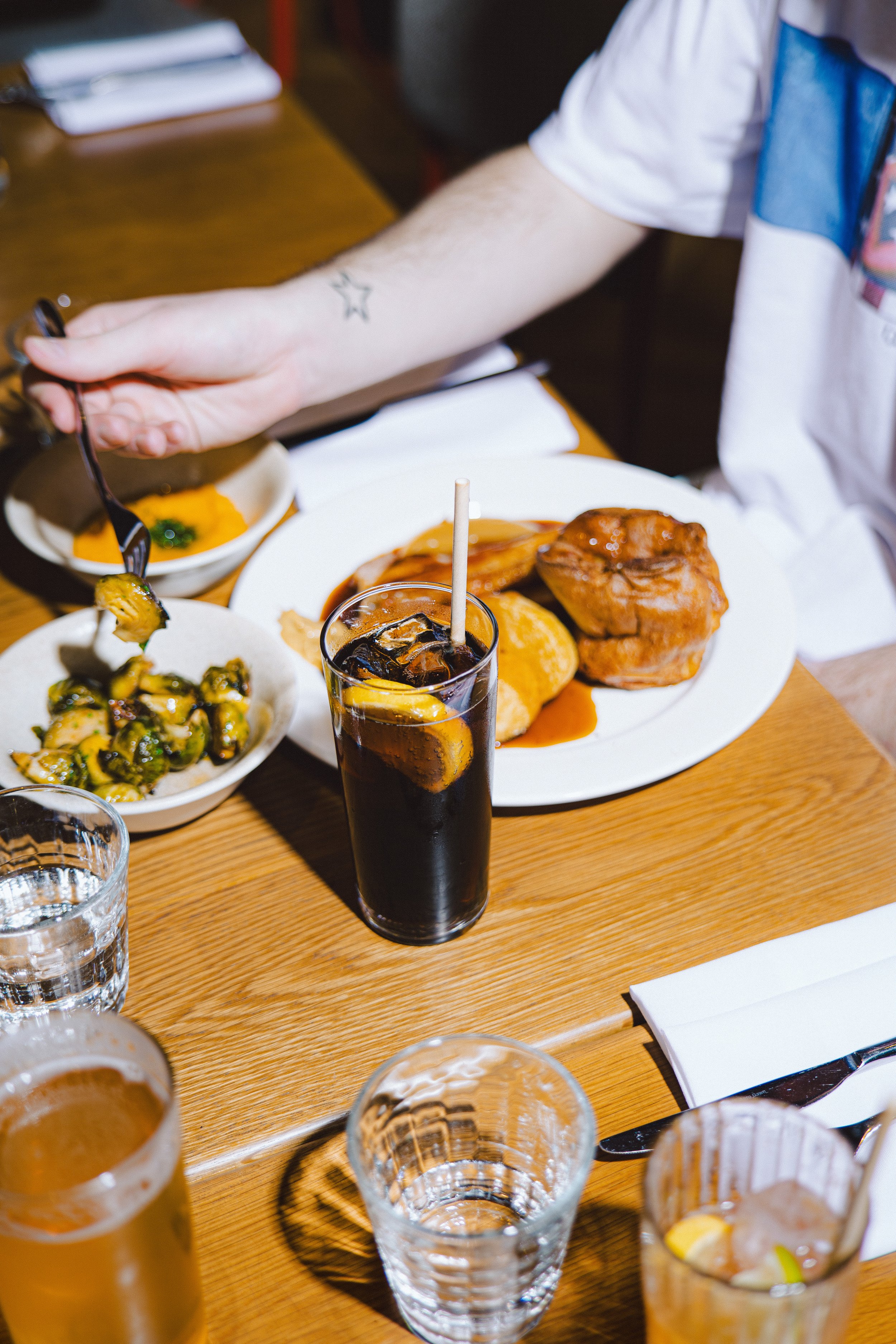 A wooden table set with various foods including a plate of meats, a bowl of Brussels sprouts, glasses of water and a dark soda with lemon, and some napkins. A person is reaching for food with a fork.