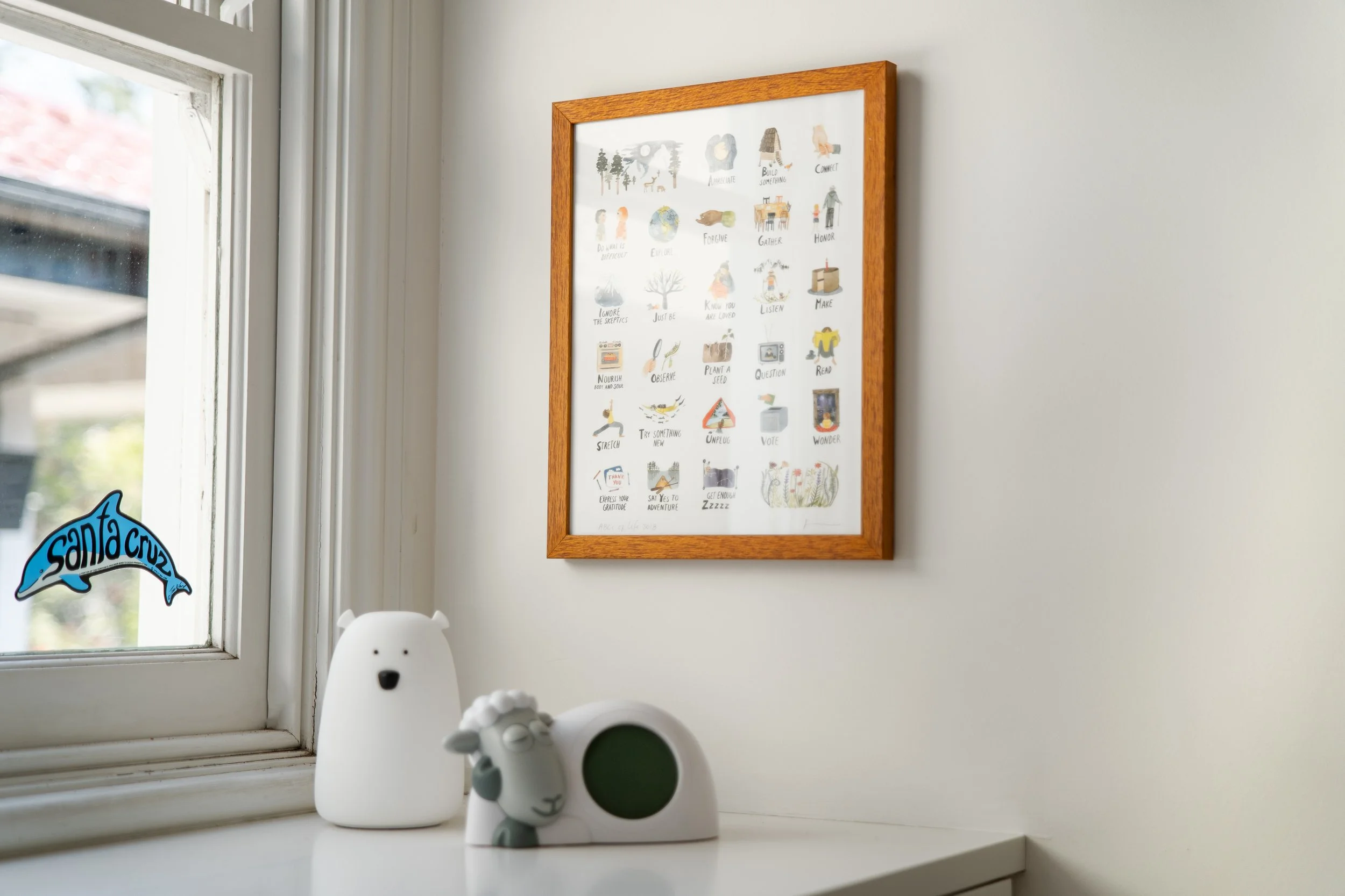 Decorative items on a windowsill, including ceramic bear and sheep figurines, with a framed watercolor art piece on a white wall behind them.