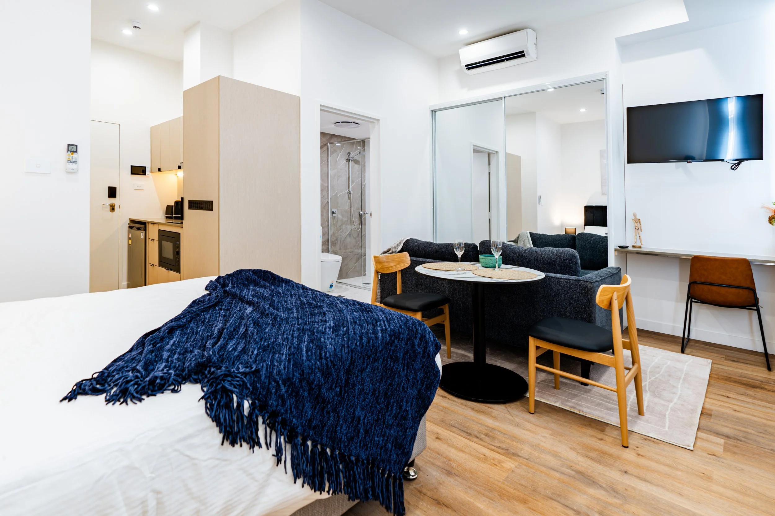 Interior of a modern studio apartment with a bed covered with a blue throw, a small kitchen area, a seating area with a round table and two chairs, a TV mounted on the wall, and a bathroom with a shower.