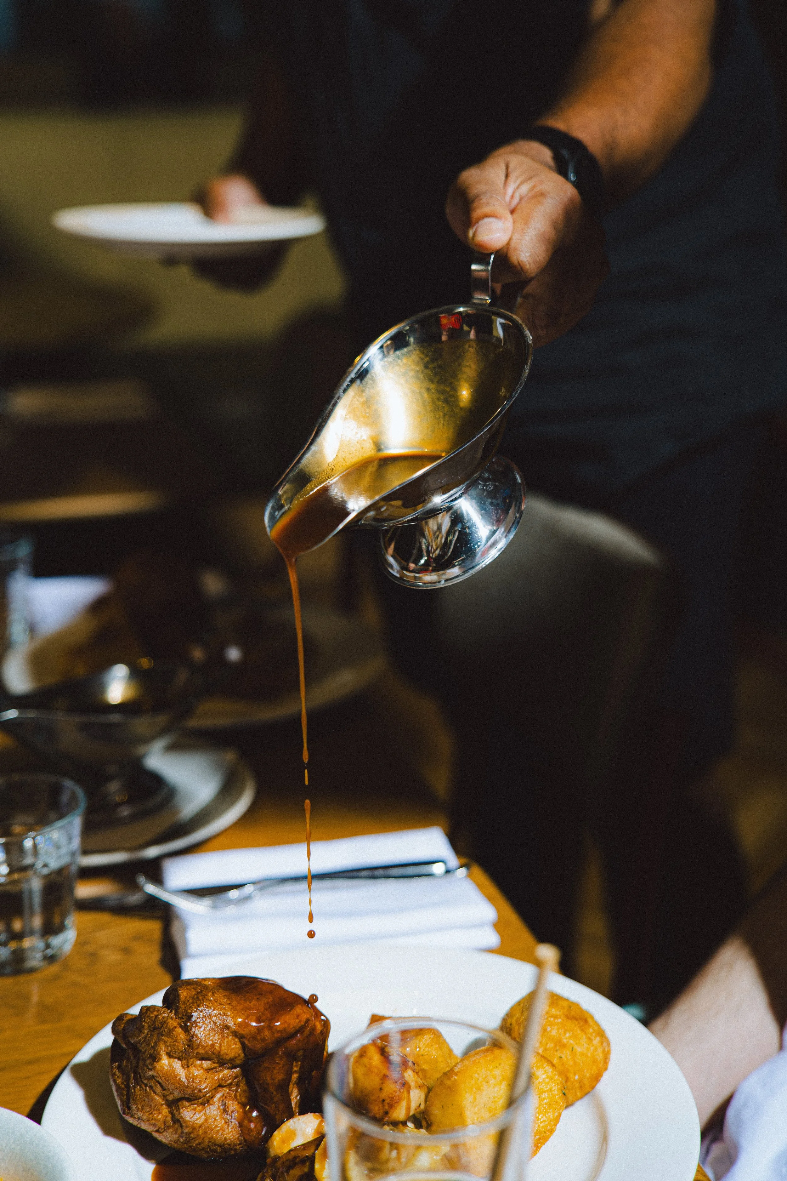 A person pouring gravy or sauce onto a plate of food including fried chicken and roasted potatoes at a dining table.
