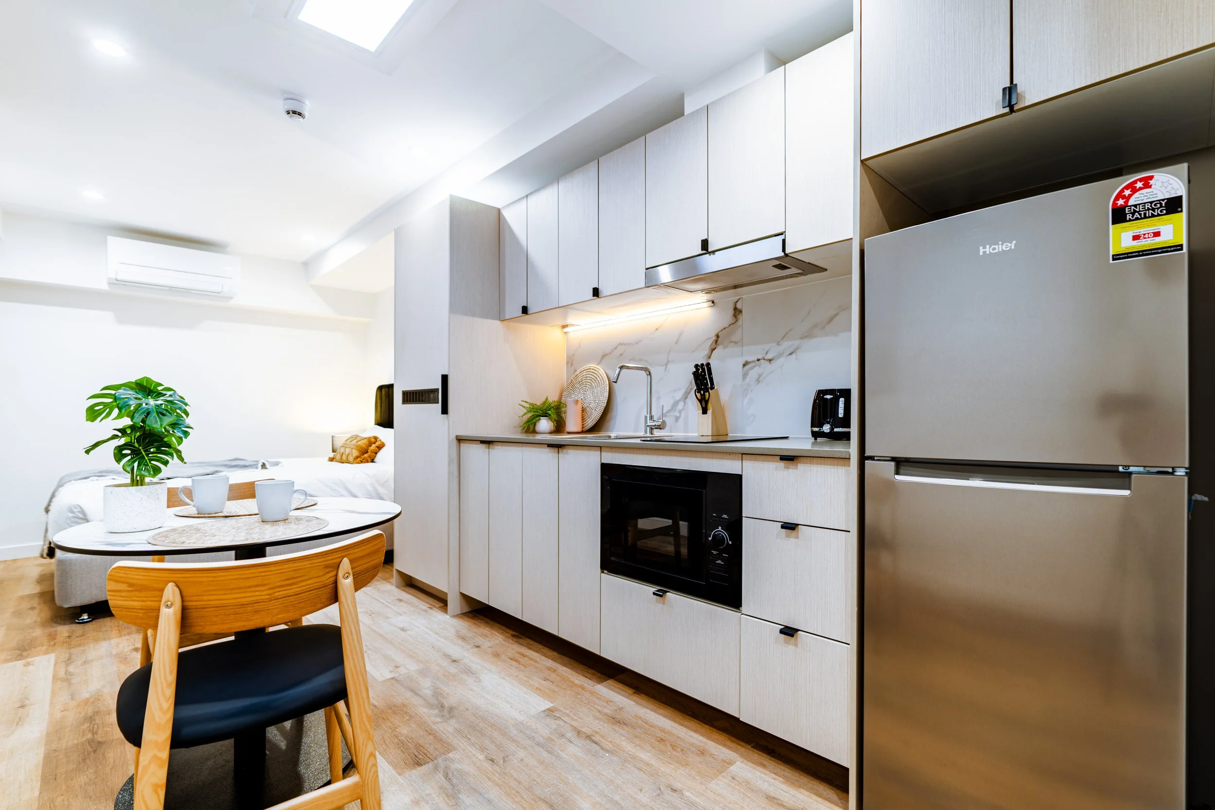 Modern studio apartment interior with kitchen area featuring a refrigerator, microwave, and countertop, next to a small dining table with chairs, and a bed in the background, decorated with plants and minimalistic decor.