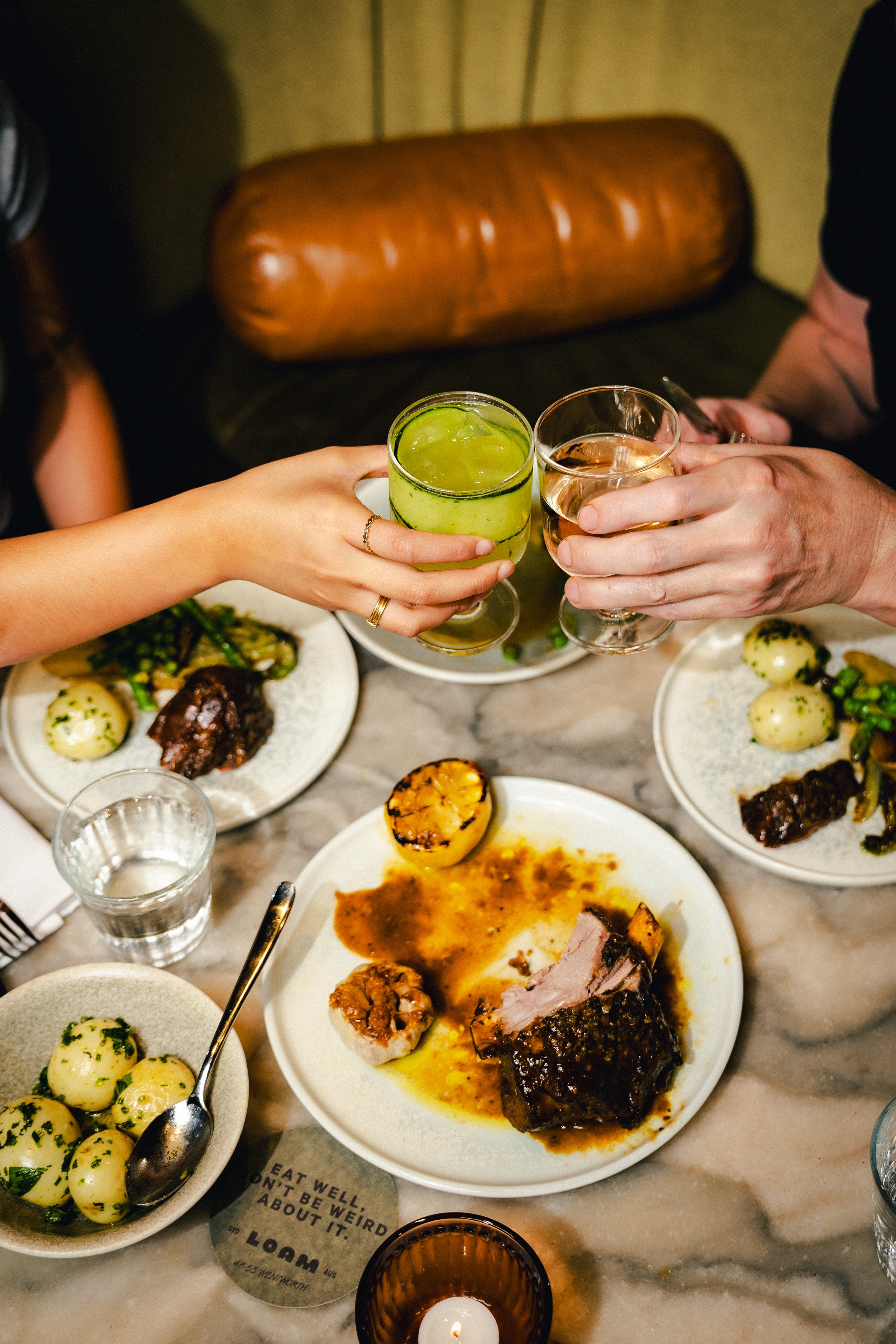 People toasting with drinks over a partially eaten meal with potatoes, grilled meat, and vegetables on a marble table.