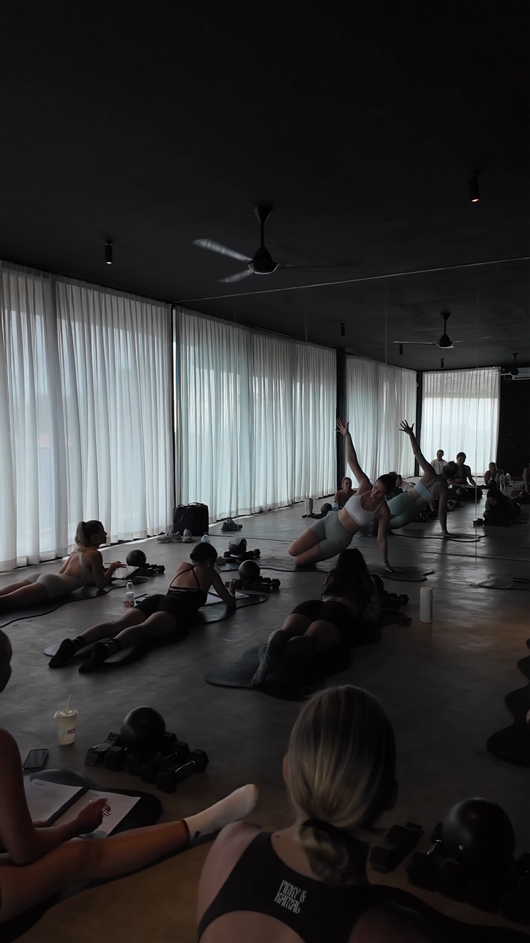 Women participating in a fitness class doing floor exercises in a dimly lit studio with large windows and sheer curtains.
