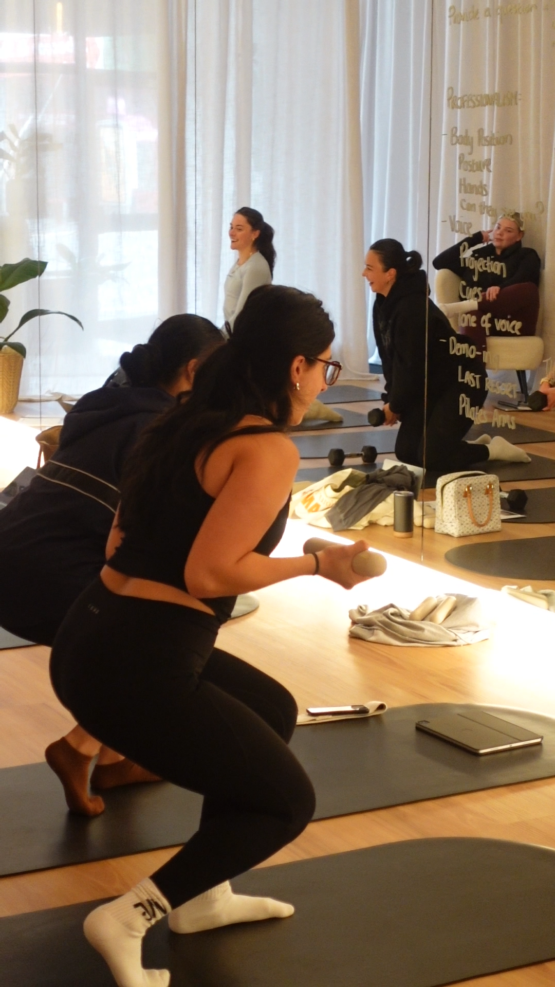A yoga class in session with several women participating, including a woman in the foreground holding a massage ball, and others seated or kneeling on yoga mats, with a large mirror and white curtains in the background.