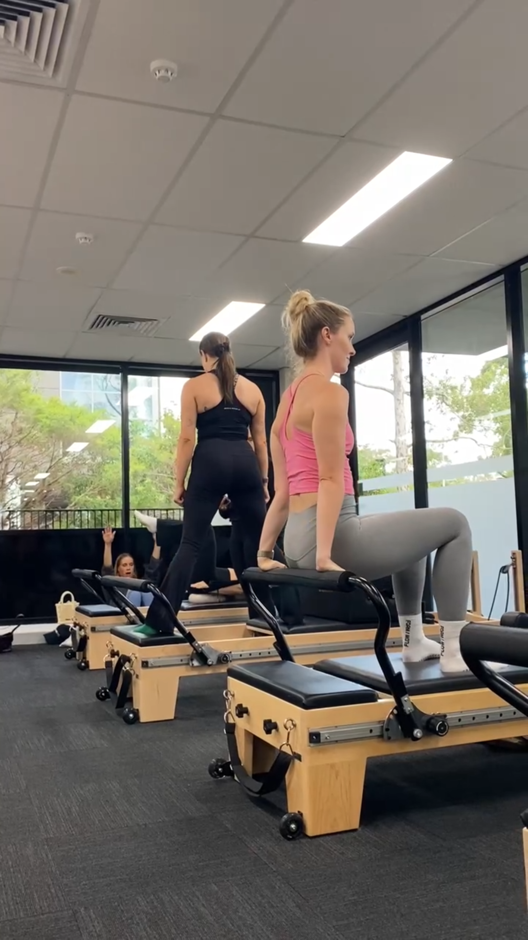 Three women working out on reformer Pilates machines in a fitness studio with large windows and natural light.