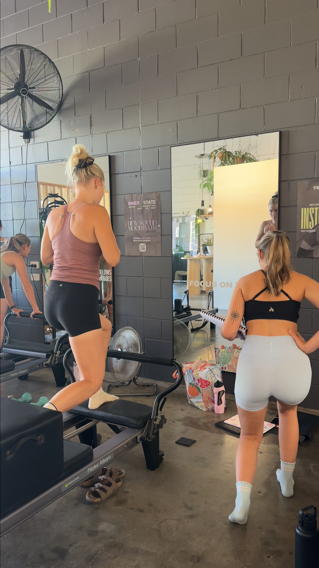 Two women exercising on Pilates reformer machines in a gym, with one woman on a reformer and another standing beside her, looking in a mirror, surrounded by workout accessories and posters on the wall.