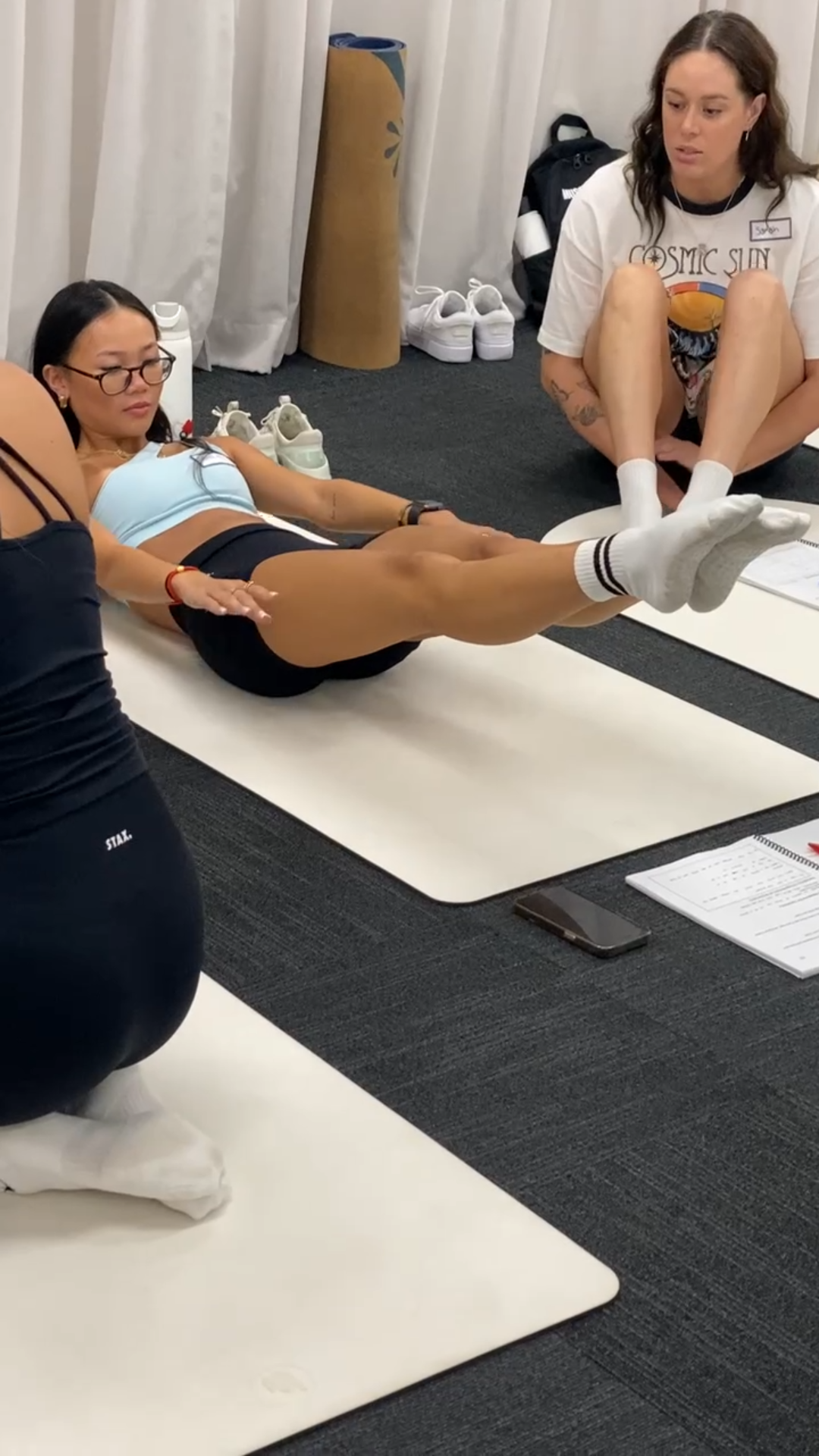 A woman in workout clothes doing leg lifts on a mat while two women assist her during a fitness or physical therapy session.