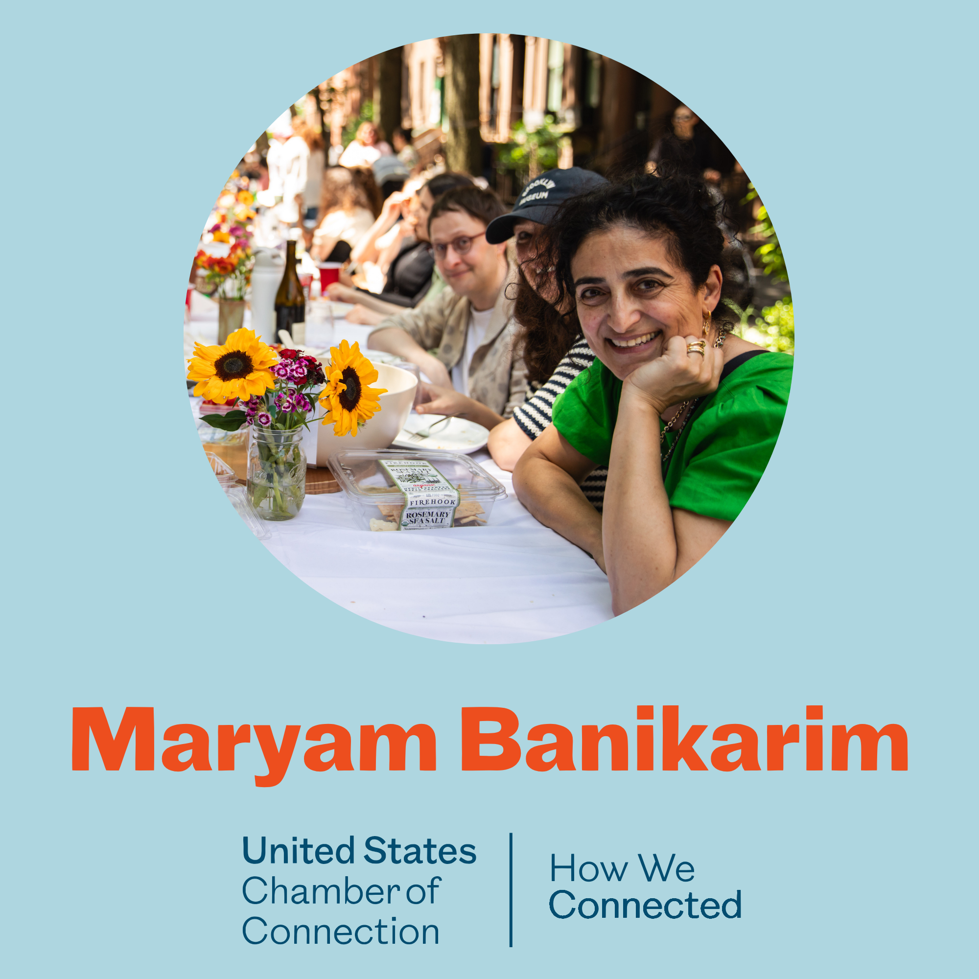 People gathered at a long outdoor table with flowers, bottles, and food in a wooded setting.