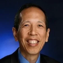 A smiling man with dark hair, wearing a blue shirt, against a dark blue background.