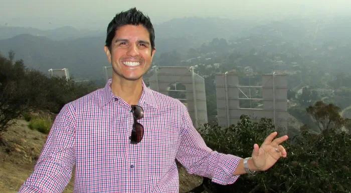Smiling man wearing a checkered shirt and sunglasses hanging from the collar, standing near the Hollywood sign on a hillside in Los Angeles with hills and fog in the background.
