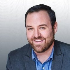 Headshot of a smiling man with dark hair and beard, wearing a navy blazer and blue shirt, against a gradient gray background.