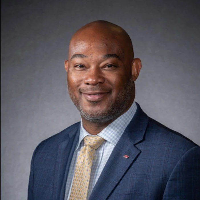 A smiling African American man wearing a blue suit, white checkered shirt, and yellow tie, against a gray background.