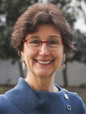 A woman with short, curly brown hair and red glasses smiling outdoors, wearing a blue sweater with a button detail.
