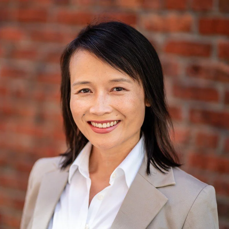 A professional woman with shoulder-length black hair, smiling, wearing a light-colored blazer over a white shirt, standing in front of a brick wall.