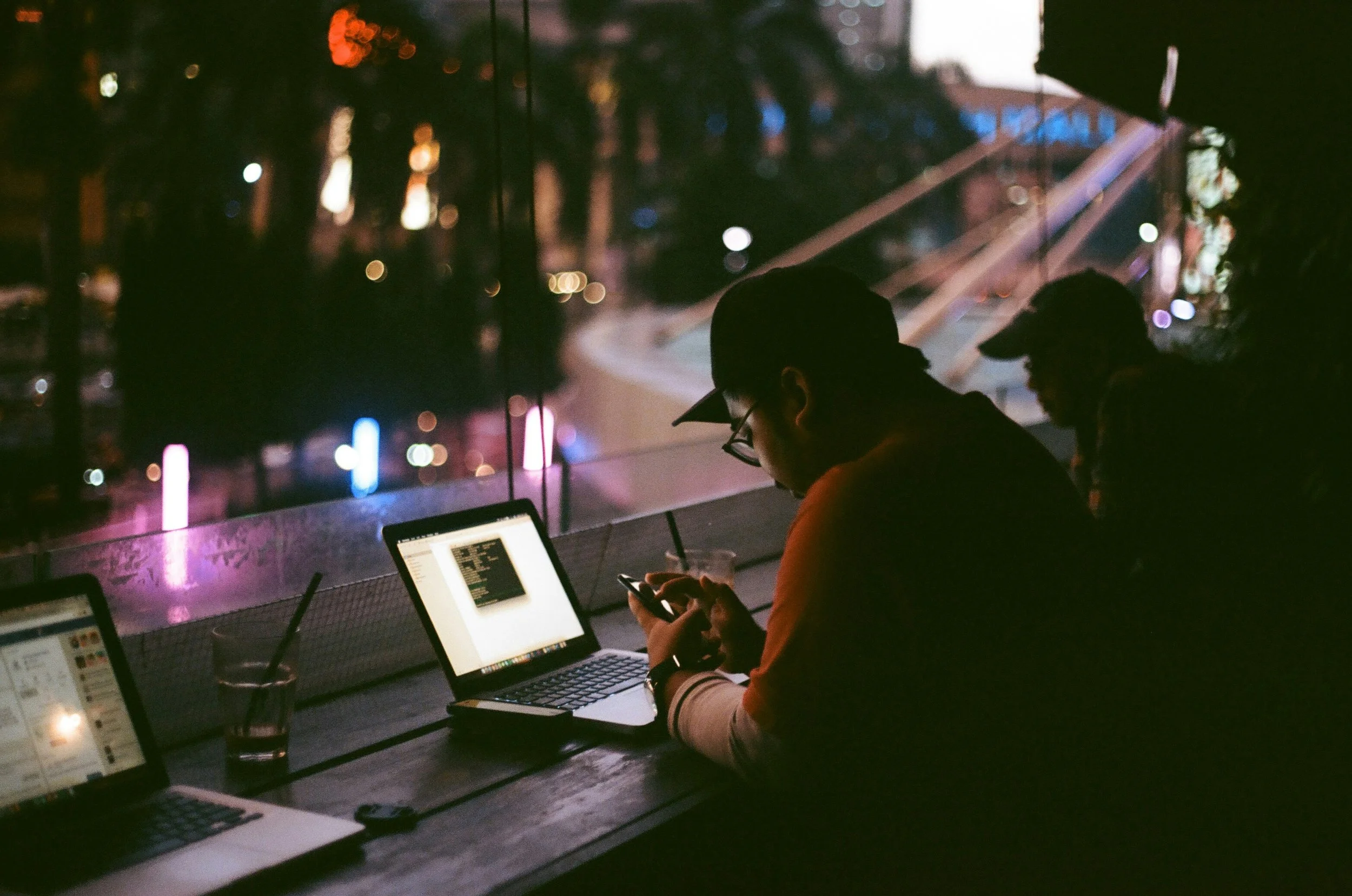 Two people sitting at a desk in a dimly lit room, working on their laptops, with a cityscape visible through the window behind them, illuminated by colorful lights.