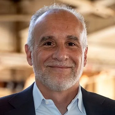 A smiling man with gray hair and beard wearing a dark blazer and light blue shirt, standing indoors with a blurred background.
