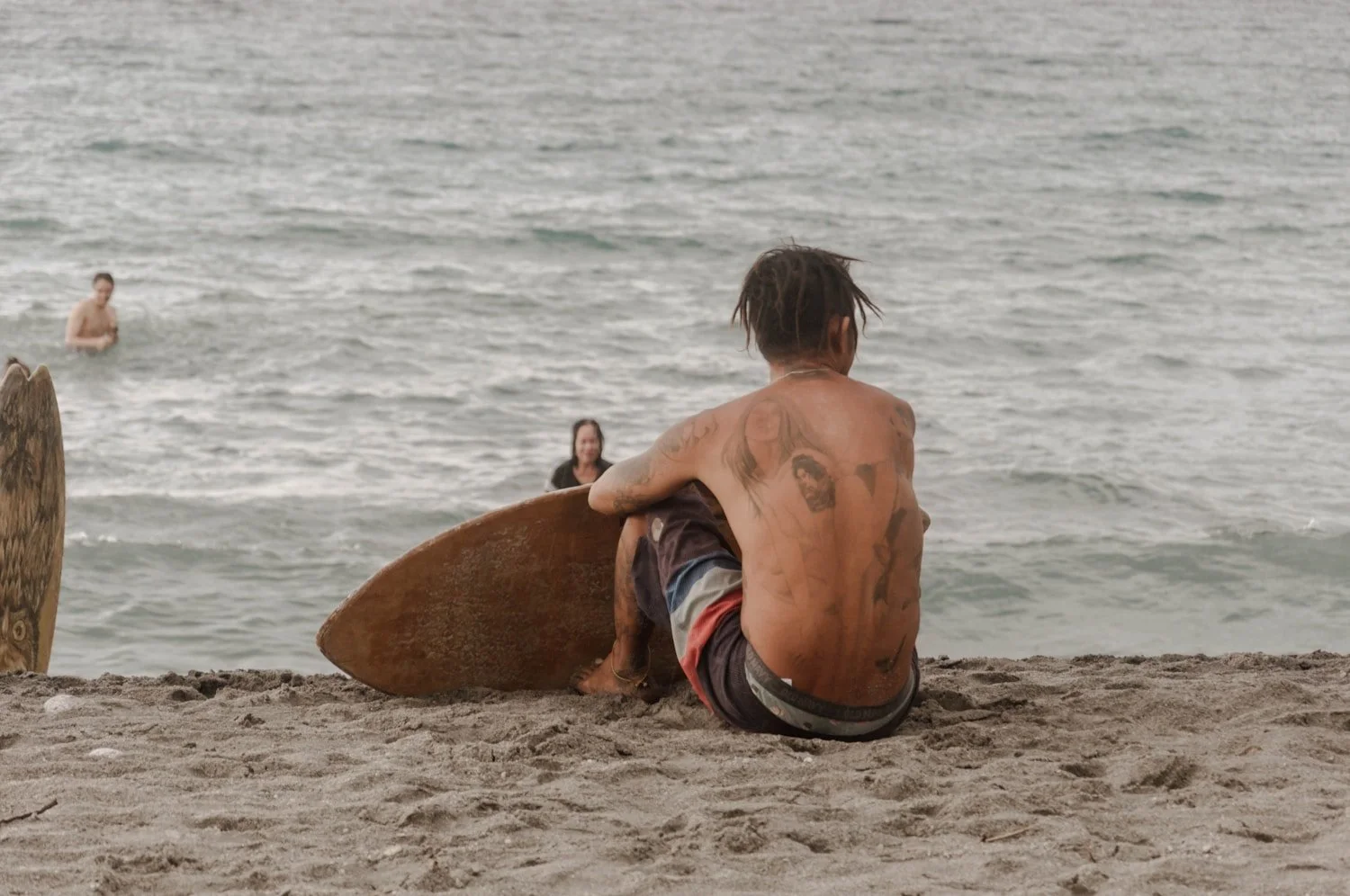 An older man sitting on the sand beside his surfboard
