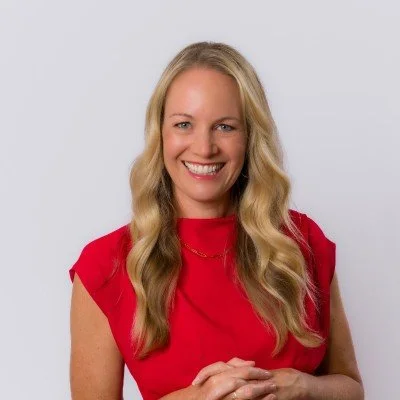 A woman with shoulder-length wavy brown hair, wearing a pink top and a multicolored beaded necklace, smiling softly against a neutral background.