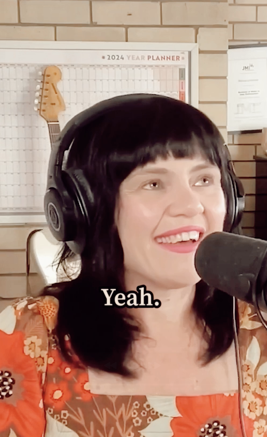 A woman with black hair and red lipstick wearing headphones and speaking into a microphone, smiling with visible teeth, in a cozy indoor setting with a 2024 planner and a brick wall in the background.