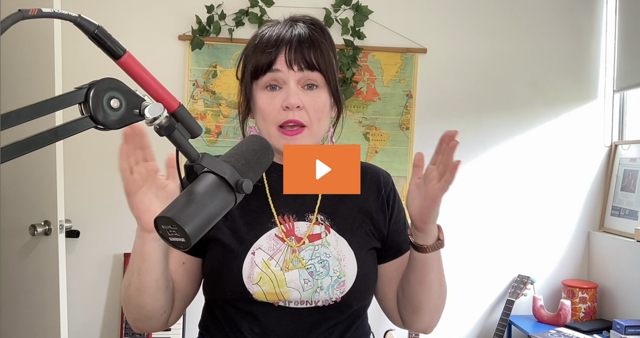 A woman with dark hair and pink lipstick speaking into a microphone in a home office or studio. She is wearing a black T-shirt with a colorful graphic design and gold jewelry. There is a world map on the wall behind her, a guitar, and some framed items nearby.
