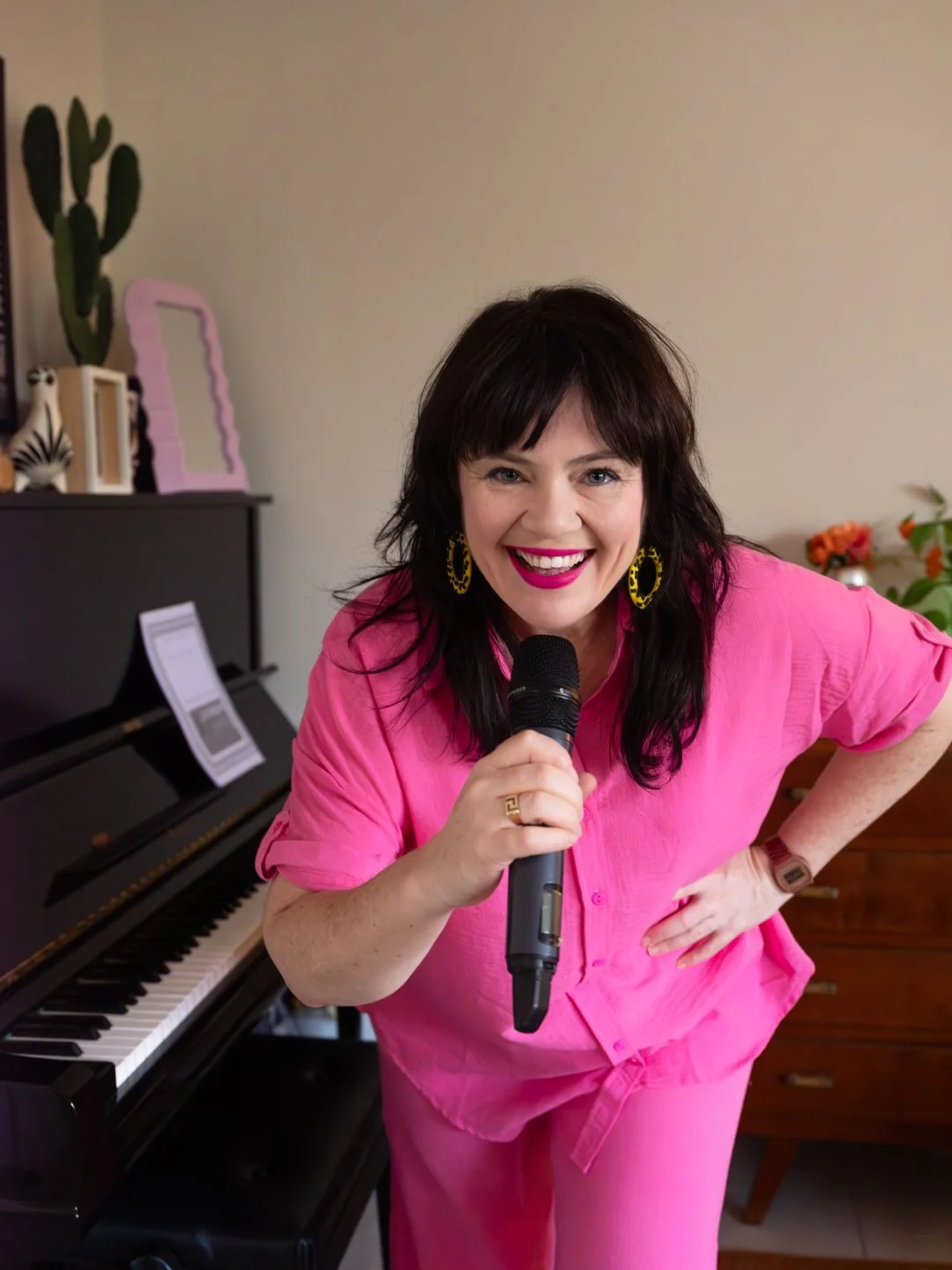 A woman with dark hair wearing a bright pink outfit and yellow earrings smiling and holding a microphone, standing beside a black piano.