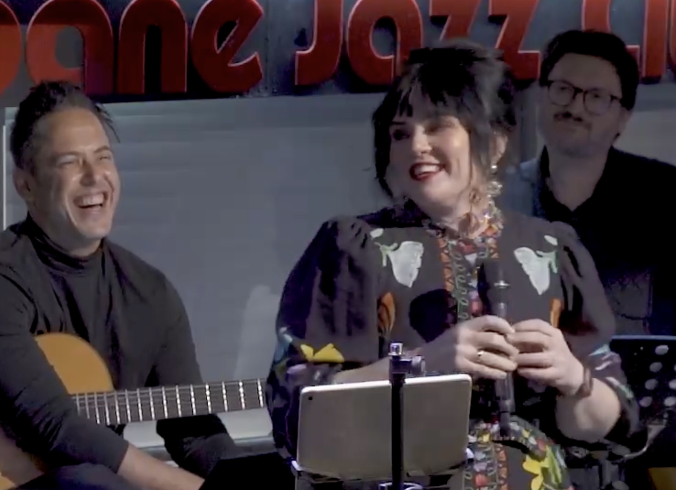 Singer Mel Lathouras smiling on stage with guitarist Eddie Gazani in a black turtleneck and bassist Samuel Vincent with an upright bass  at Brisbane Jazz Club. Mel is holding a mic, with Gorman folklore printed dress with 1960's up-do.