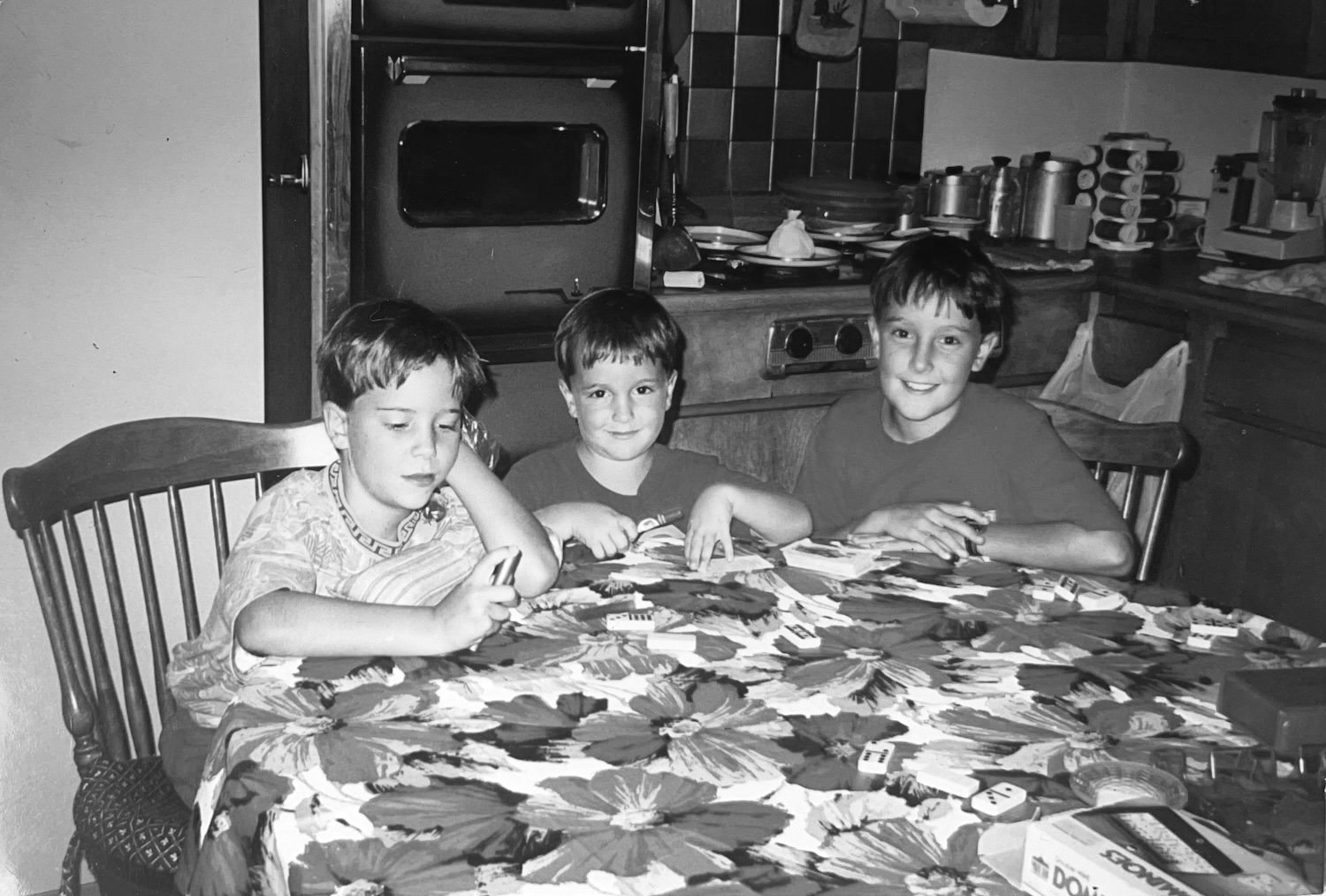 Three young boys sitting at a table in a kitchen, playing a game with small tiles. The table has a floral tablecloth. The kitchen has various pots, pans, and kitchen items in the background.