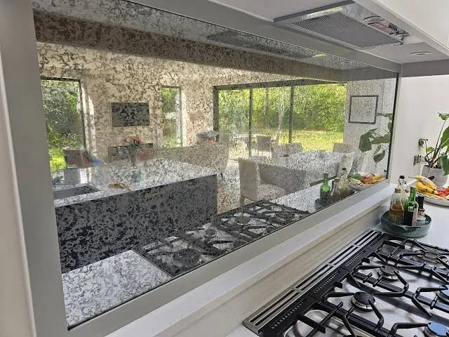 View of a modern kitchen with a granite countertop, stove, and a glass wall through which a dining area and green outdoor scenery are visible.