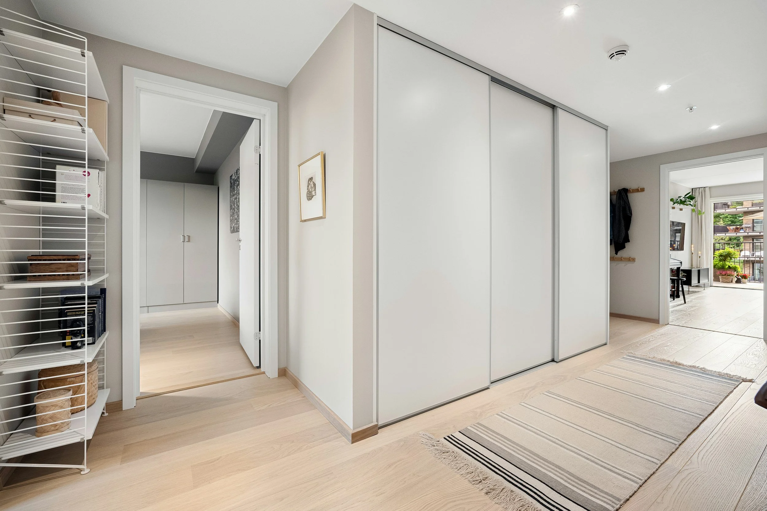 Interior view of a modern apartment with light wood flooring, a beige and white color scheme, a wardrobe with sliding doors, a shelf with books and storage baskets, and large windows with a balcony showing outdoor greenery.