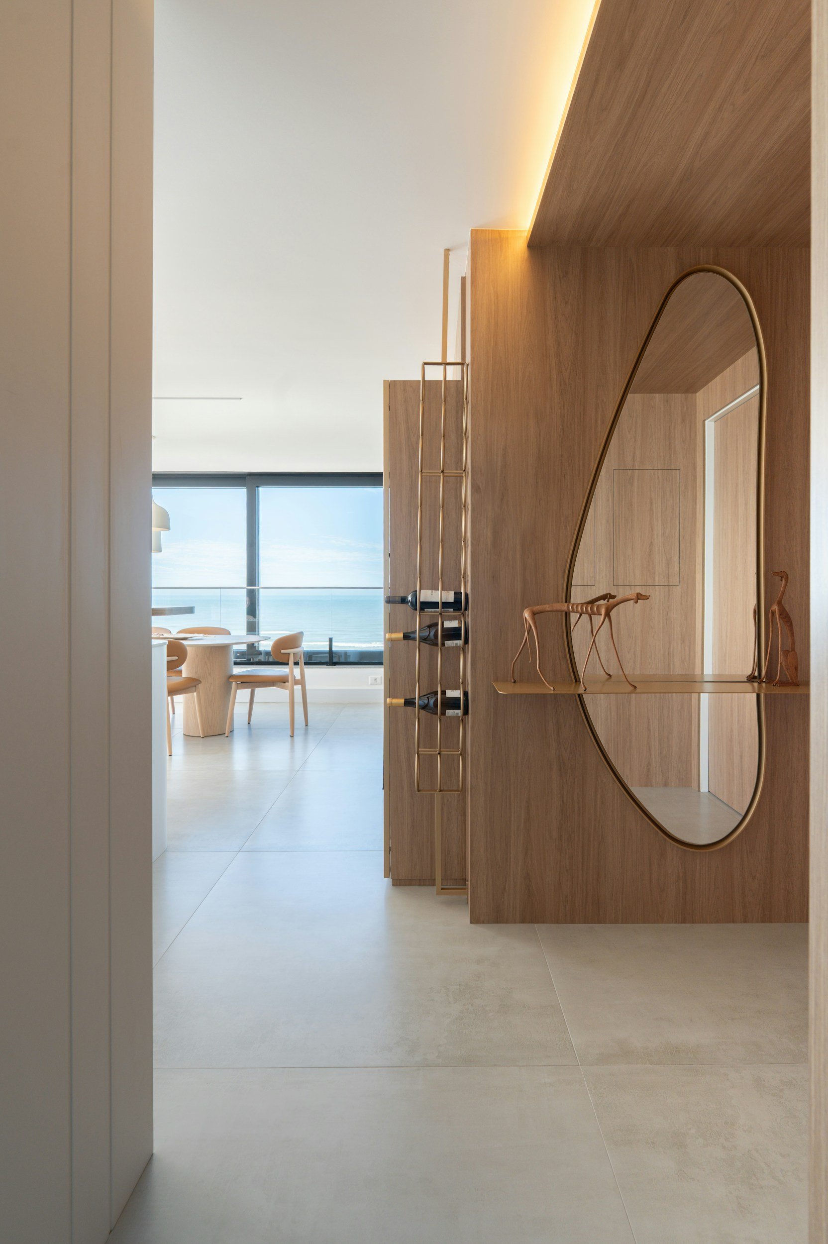 Modern interior of a room with a wooden partition featuring a circular mirror and decorative sculptures, with a view of a bright living area and large windows overlooking a body of water.