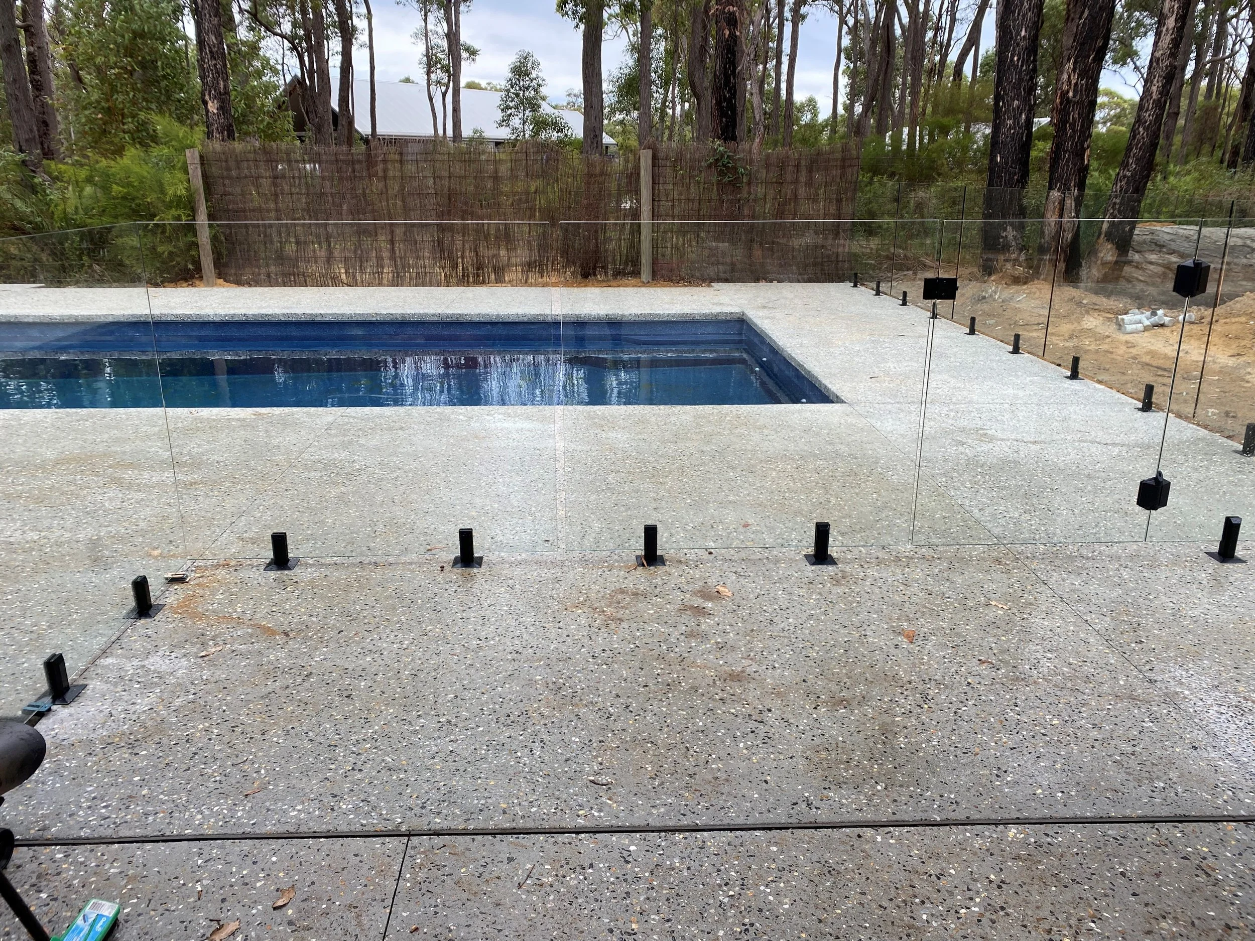 A swimming pool with a safety fence surrounding it, set on a concrete patio area. In the background, there are trees and a fence.