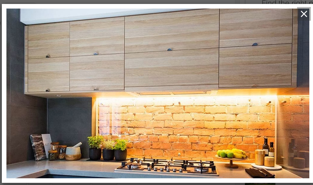 Modern kitchen with a brick backsplash illuminated by under-cabinet lighting, wooden cabinets, and a countertop stove. There are small potted plants, jars, and a fruit bowl on the counter.