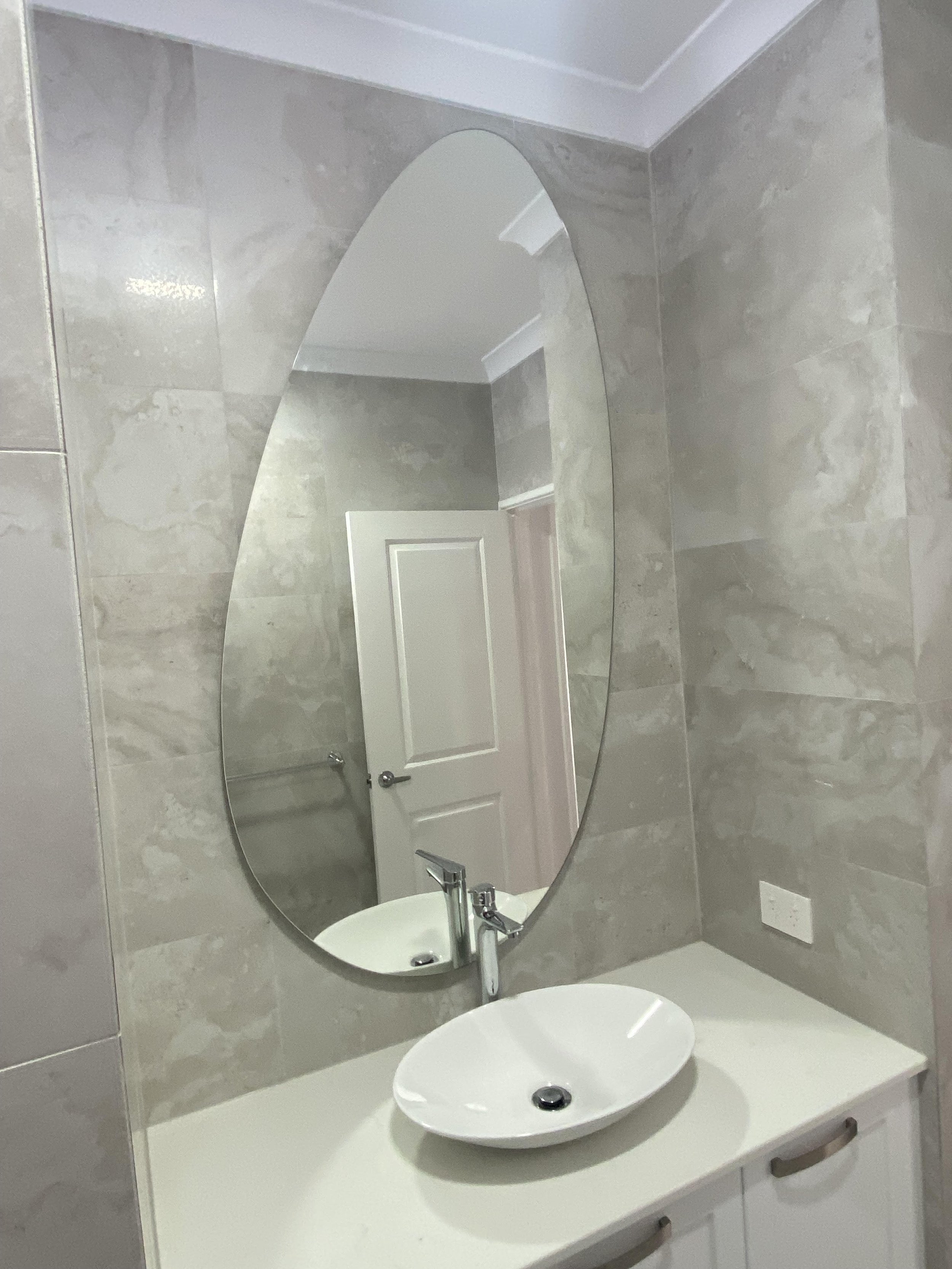 Modern bathroom with a large oval mirror, a white vessel sink with a chrome faucet, marble tile walls, and white cabinetry.