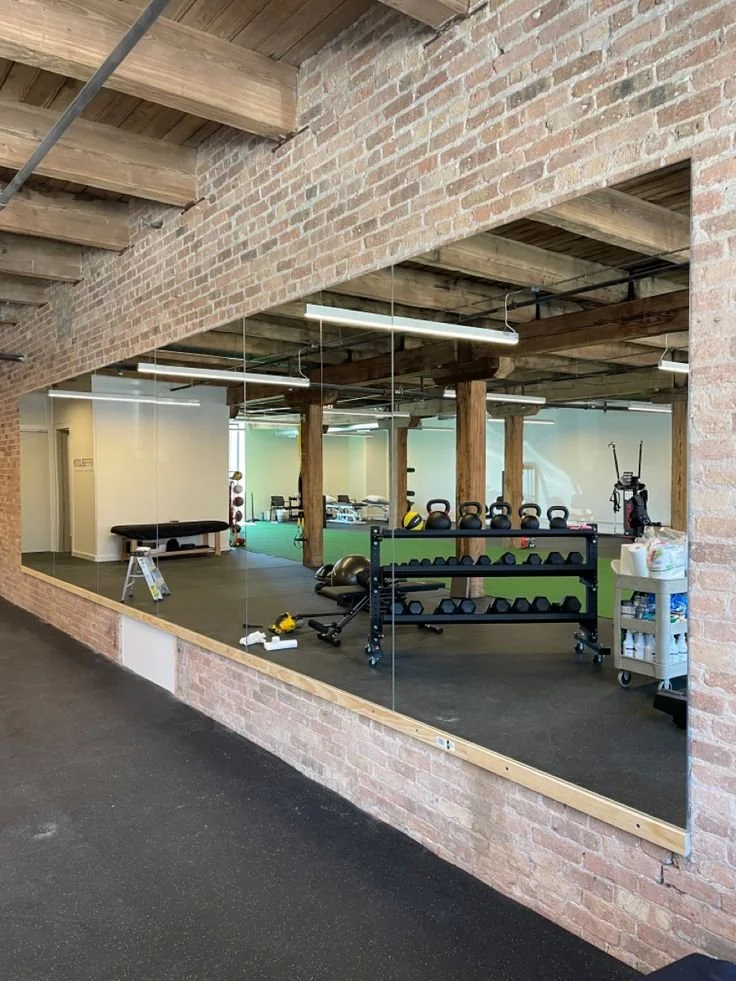 View of a gym seen through a large mirror on a brick wall, featuring kettlebells, medicine balls, and exercise equipment.