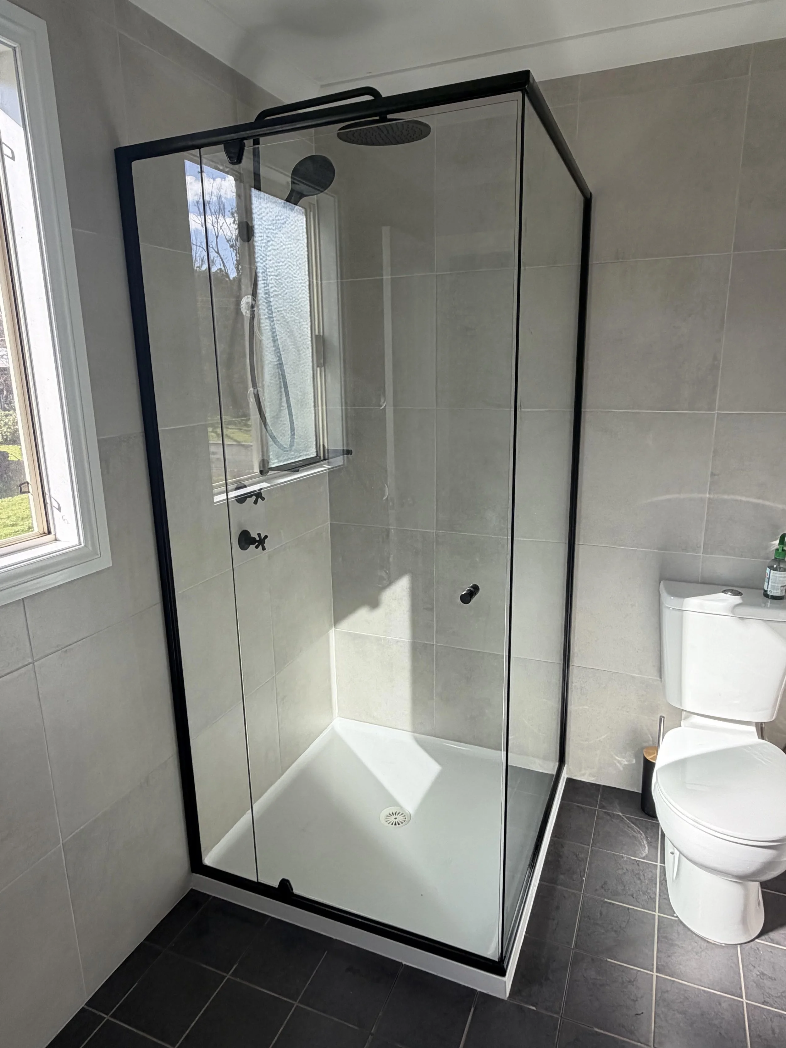 Bathroom shower with glass enclosure, overhead rainfall showerhead, handheld shower, gray tiled walls, a window, and a toilet nearby.