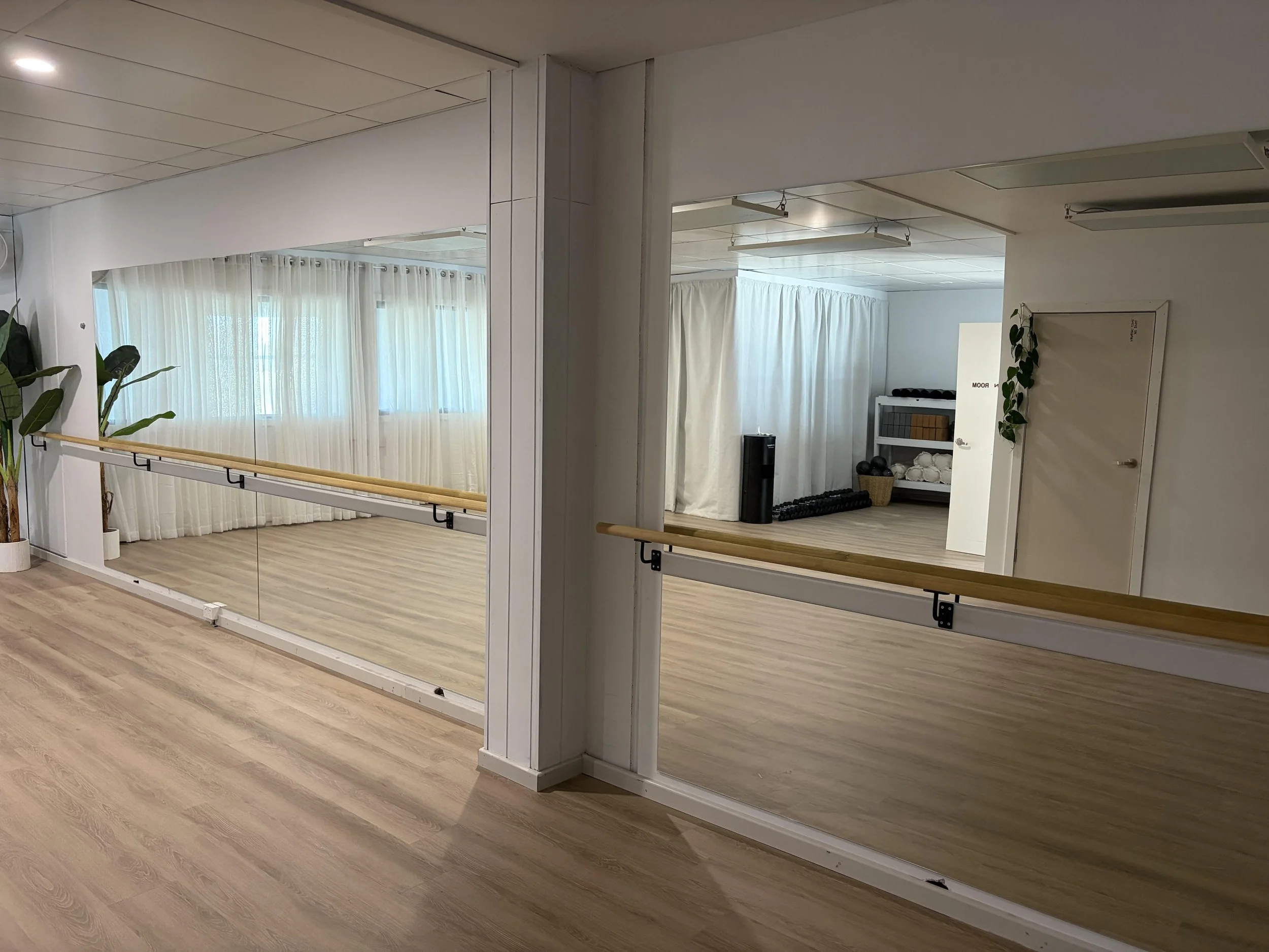 A mirror and a ballet barre in a dance studio, with white curtains by the windows, gym equipment, and wooden flooring.