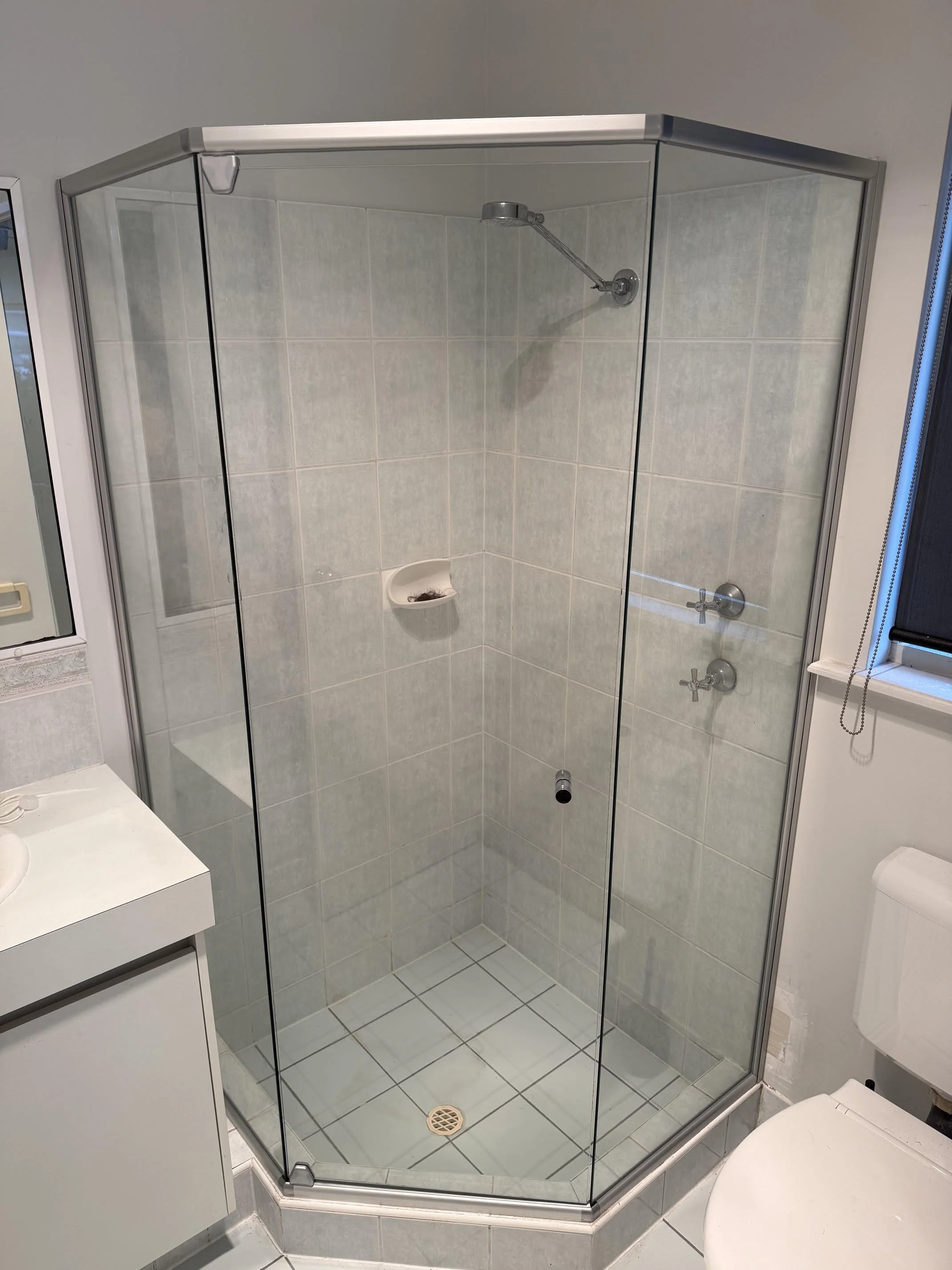 Enclosed glass shower with light-colored tiled walls, a showerhead, two control knobs, a small built-in soap dish, and a wooden door.